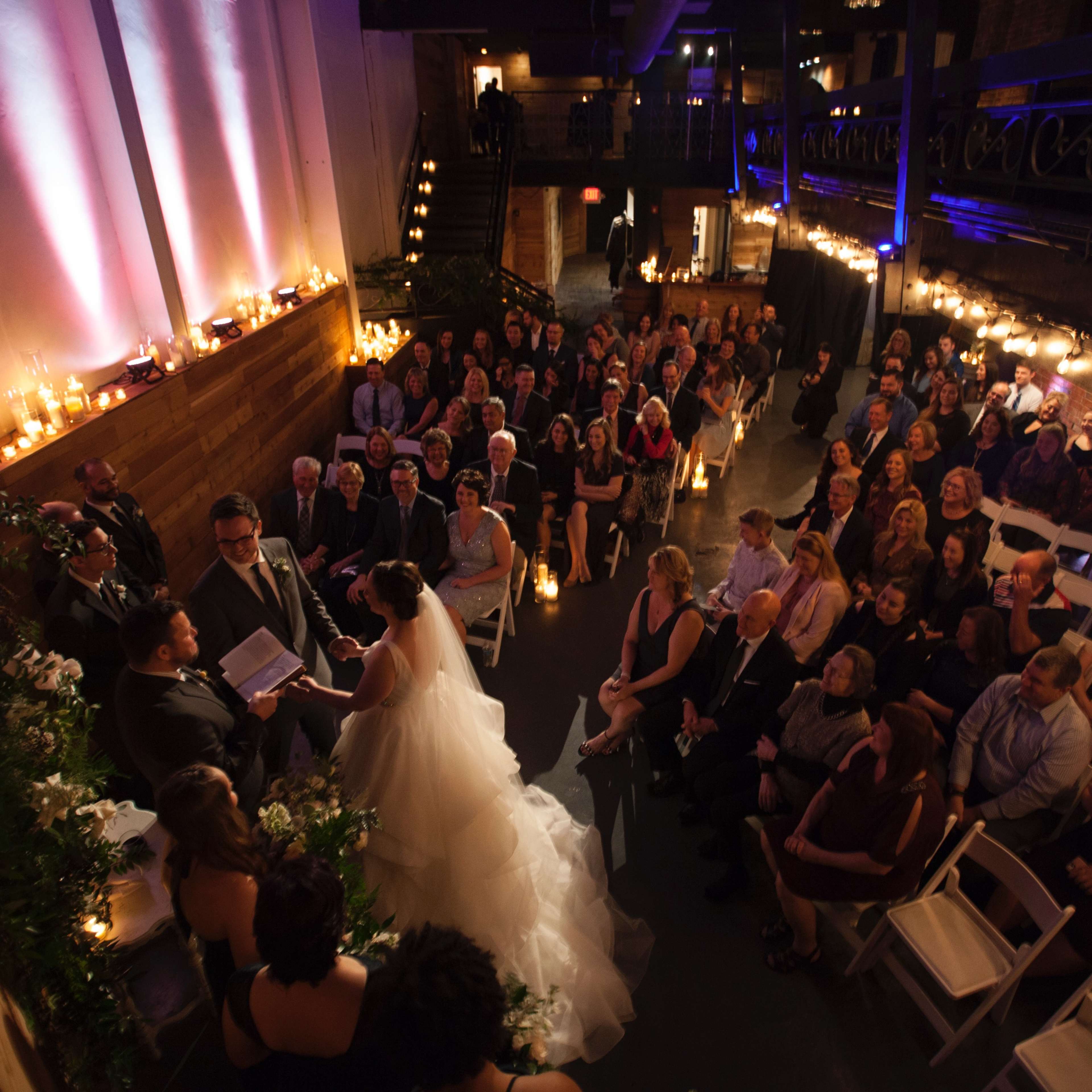 A bride and groom stand at the altar while guests seated in rows observe the wedding ceremony in a warmly lit venue.