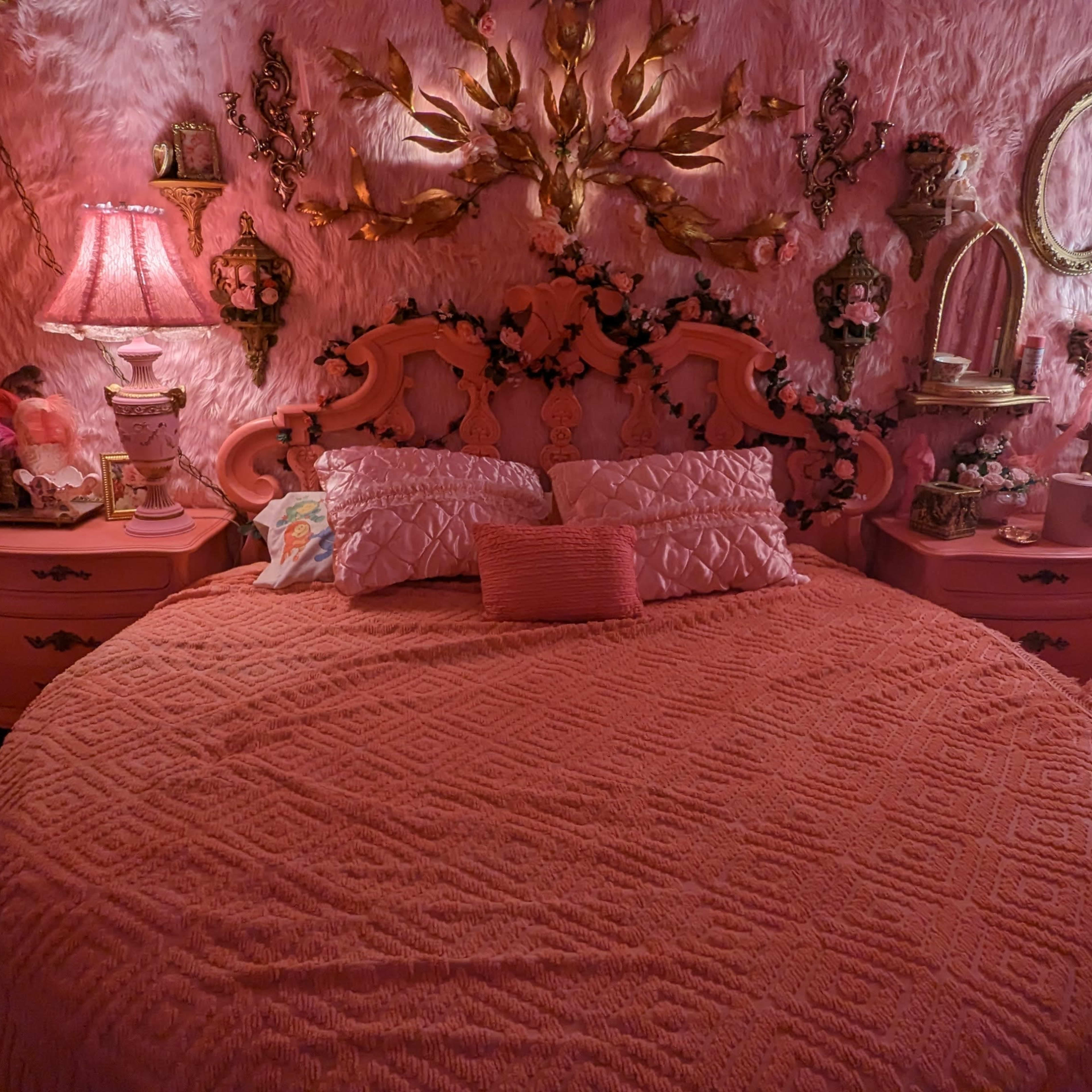 The bedroom features a round bed with a textured pink coverlet, surrounded by walls adorned with pink faux fur and decorative mirrors, lamps, and wall art.