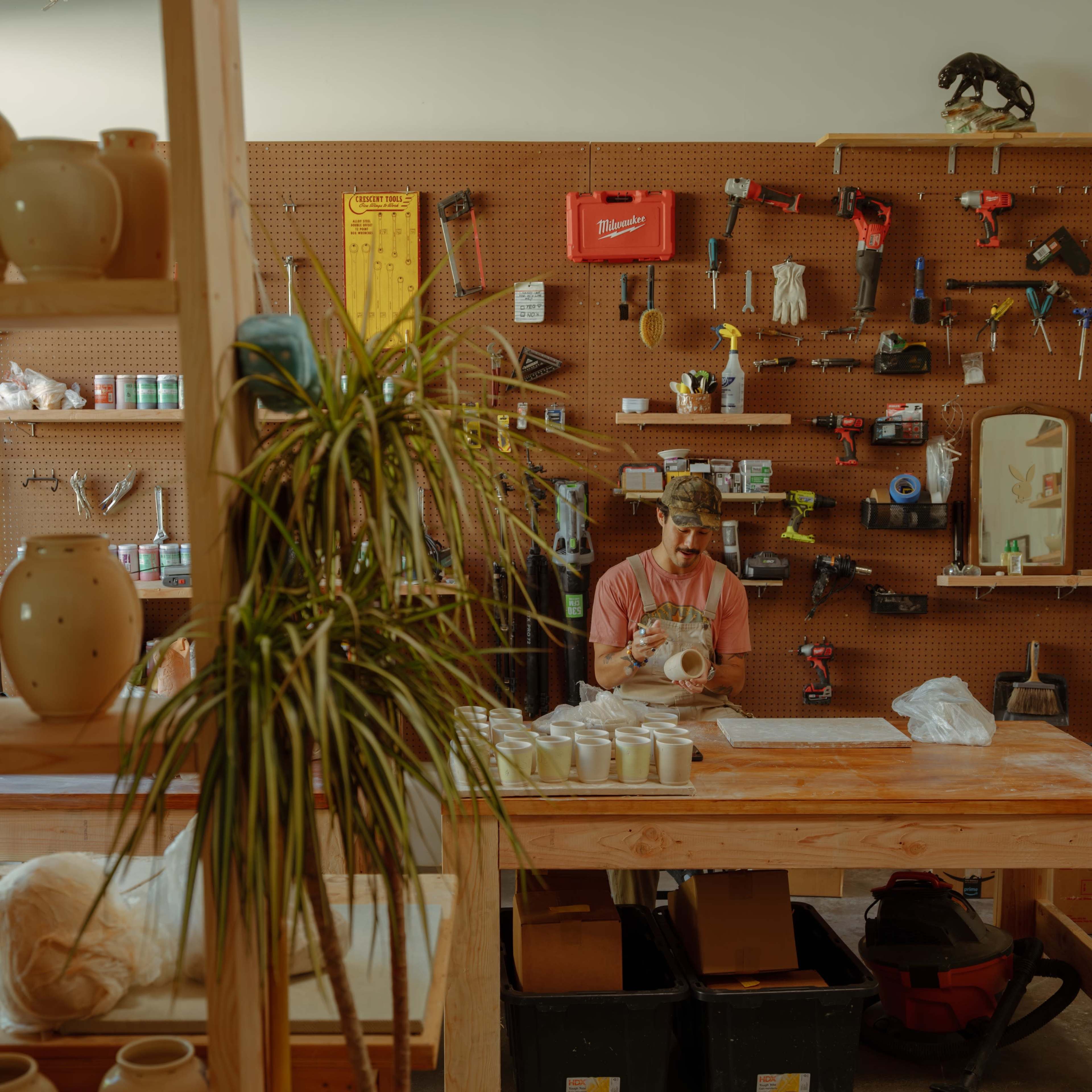 A person is seated at a wooden table in a workshop, focused on shaping a ceramic piece, surrounded by various tools and pottery on shelves.