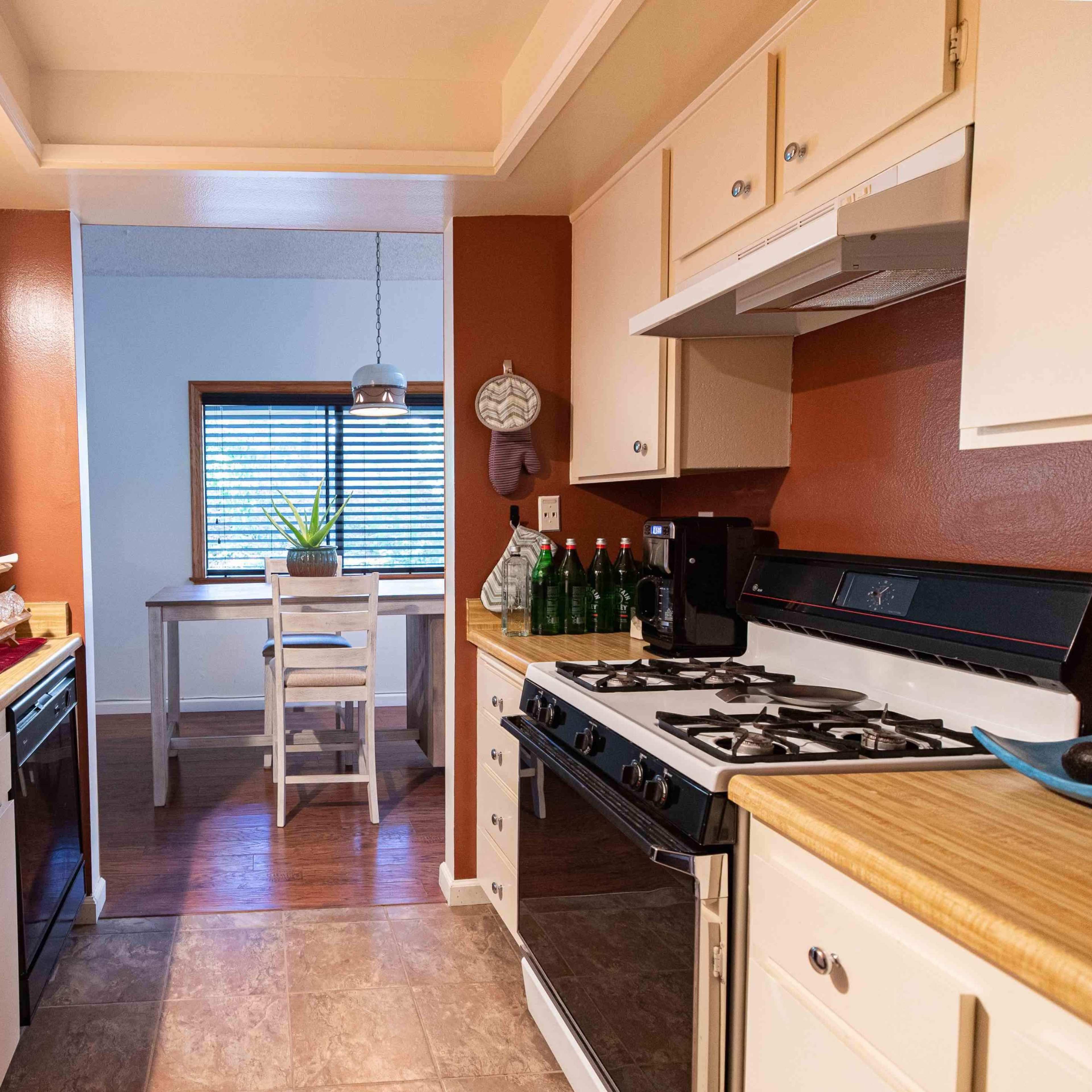 A compact kitchen with light-colored cabinets, a sink, a stove, and a small dining area in the background illuminated by a window.
