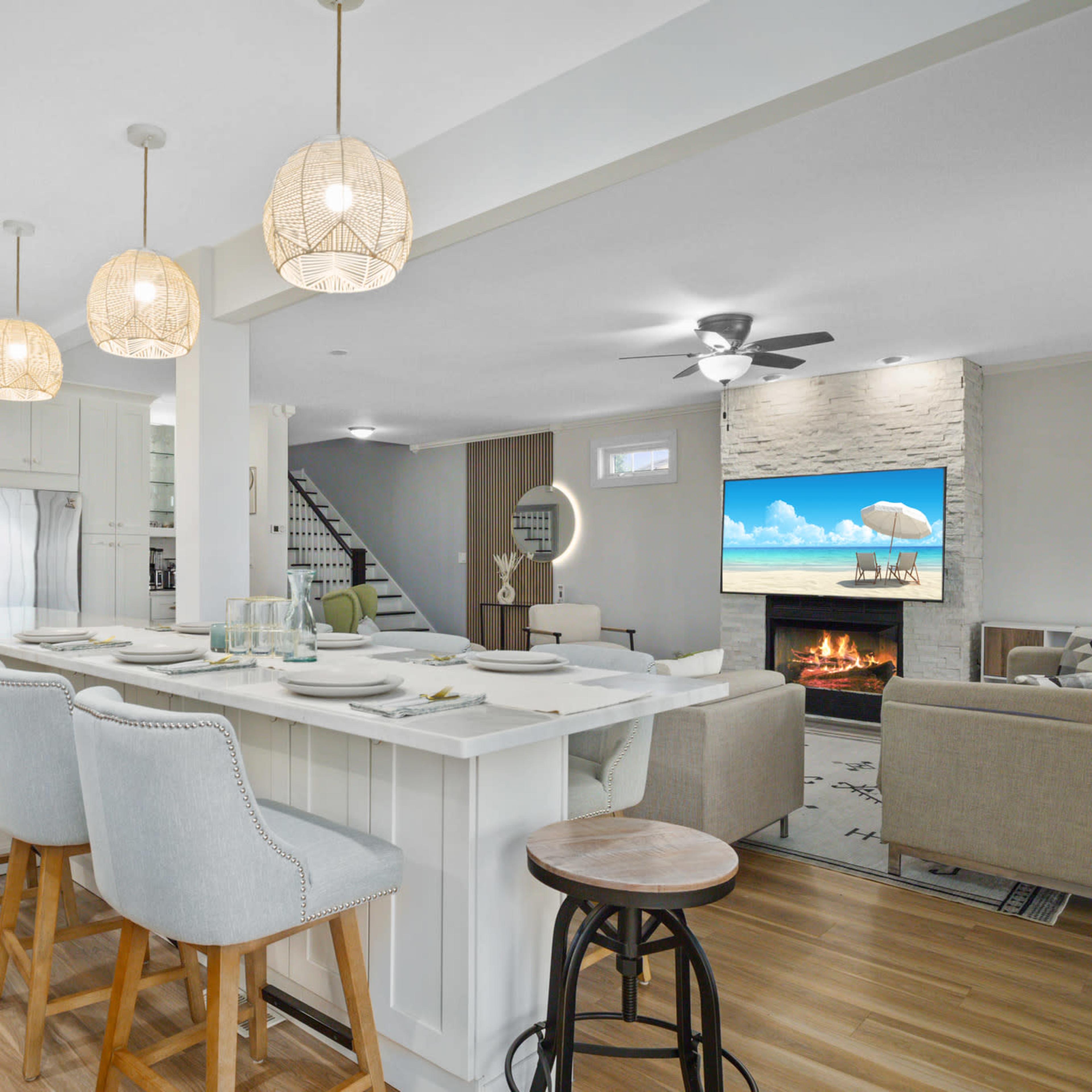 A modern kitchen features a large island with bar stools, pendant lights, and views of an adjacent living area with a fireplace and a television.