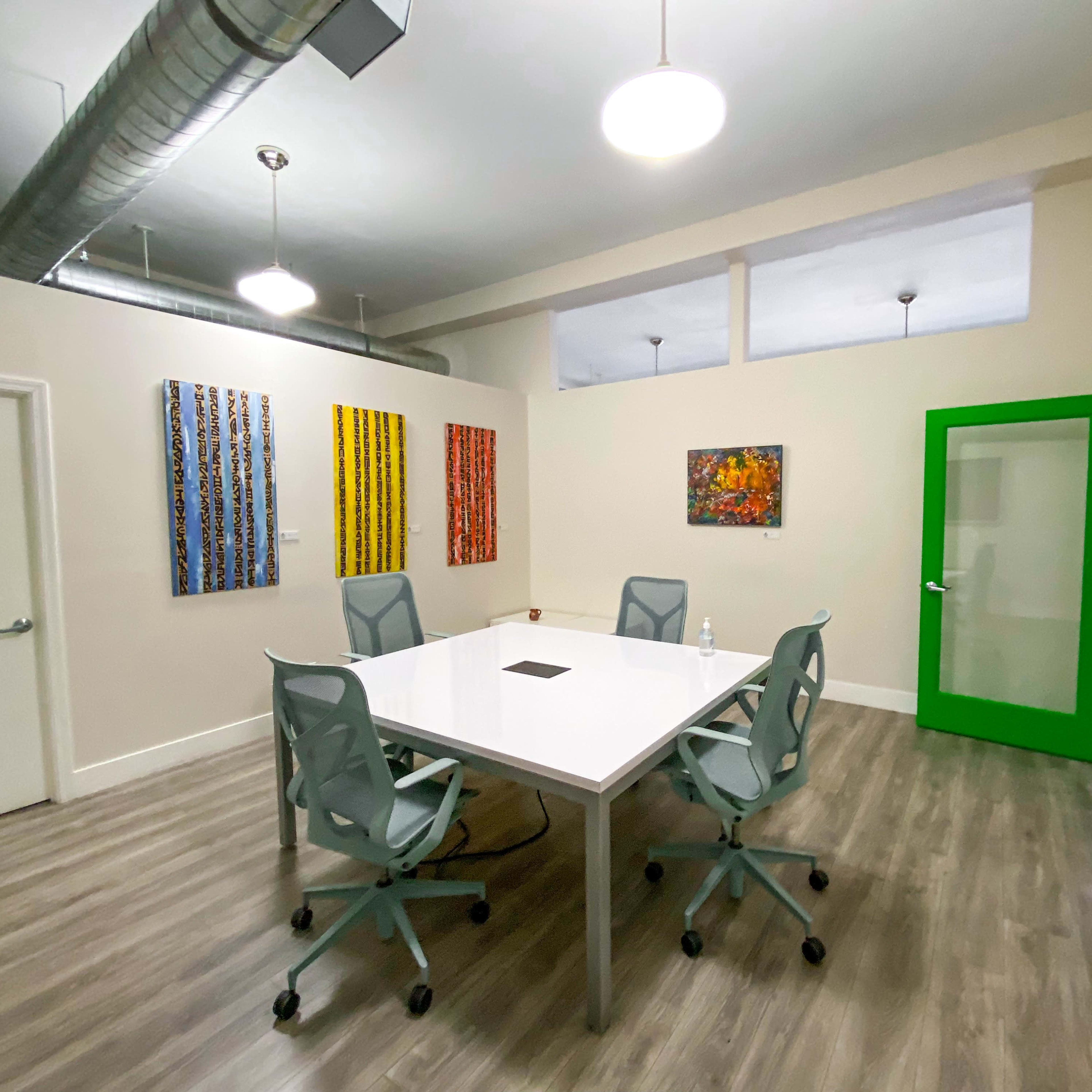 A modern conference room features a large white table surrounded by gray chairs, with colorful artwork displayed on the walls and a green glass door leading to another space.