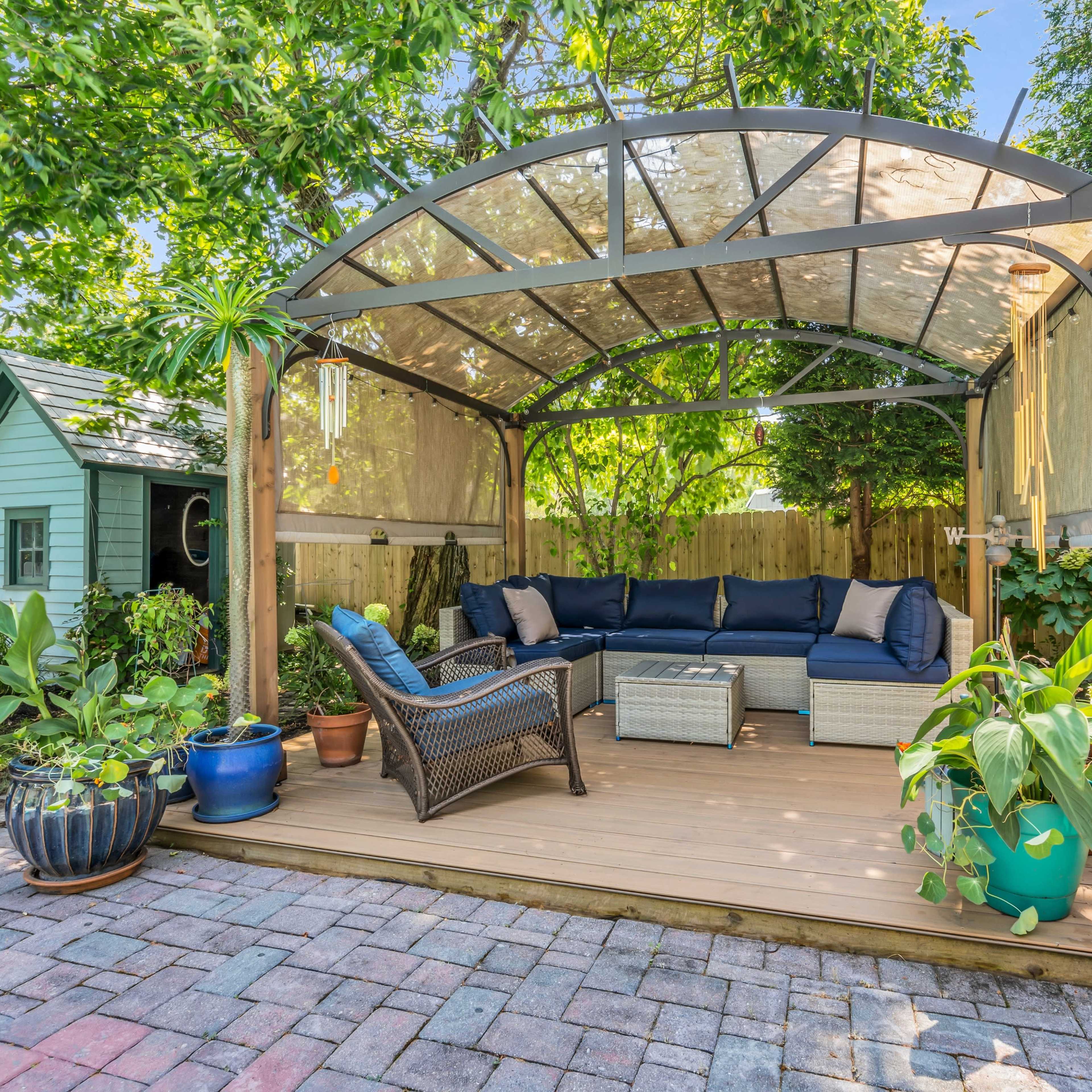 A covered outdoor seating area with a sectional sofa, coffee table, and various potted plants is surrounded by greenery and a small garden shed.