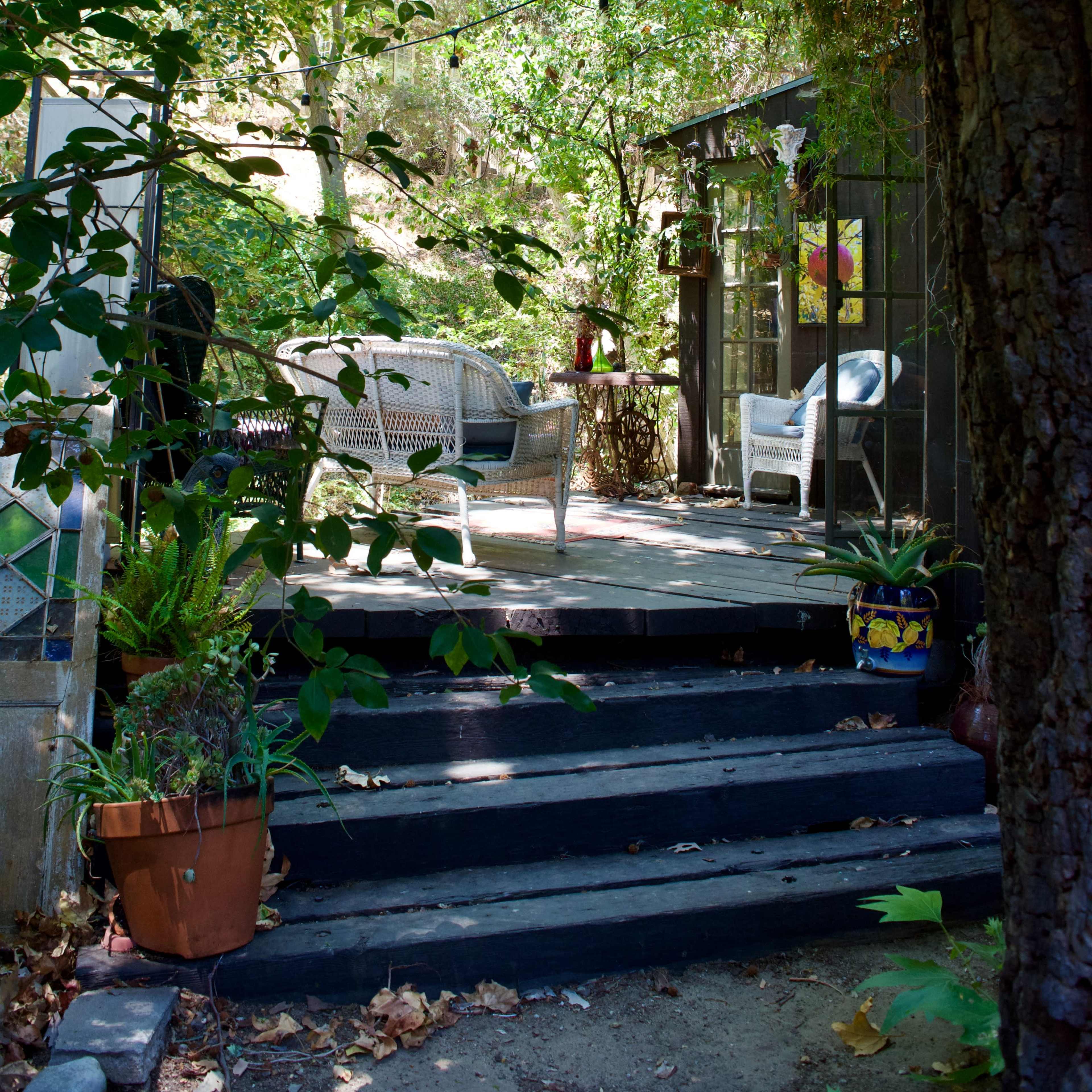A set of wooden steps leads to a shaded outdoor seating area surrounded by lush greenery and a rustic building.
