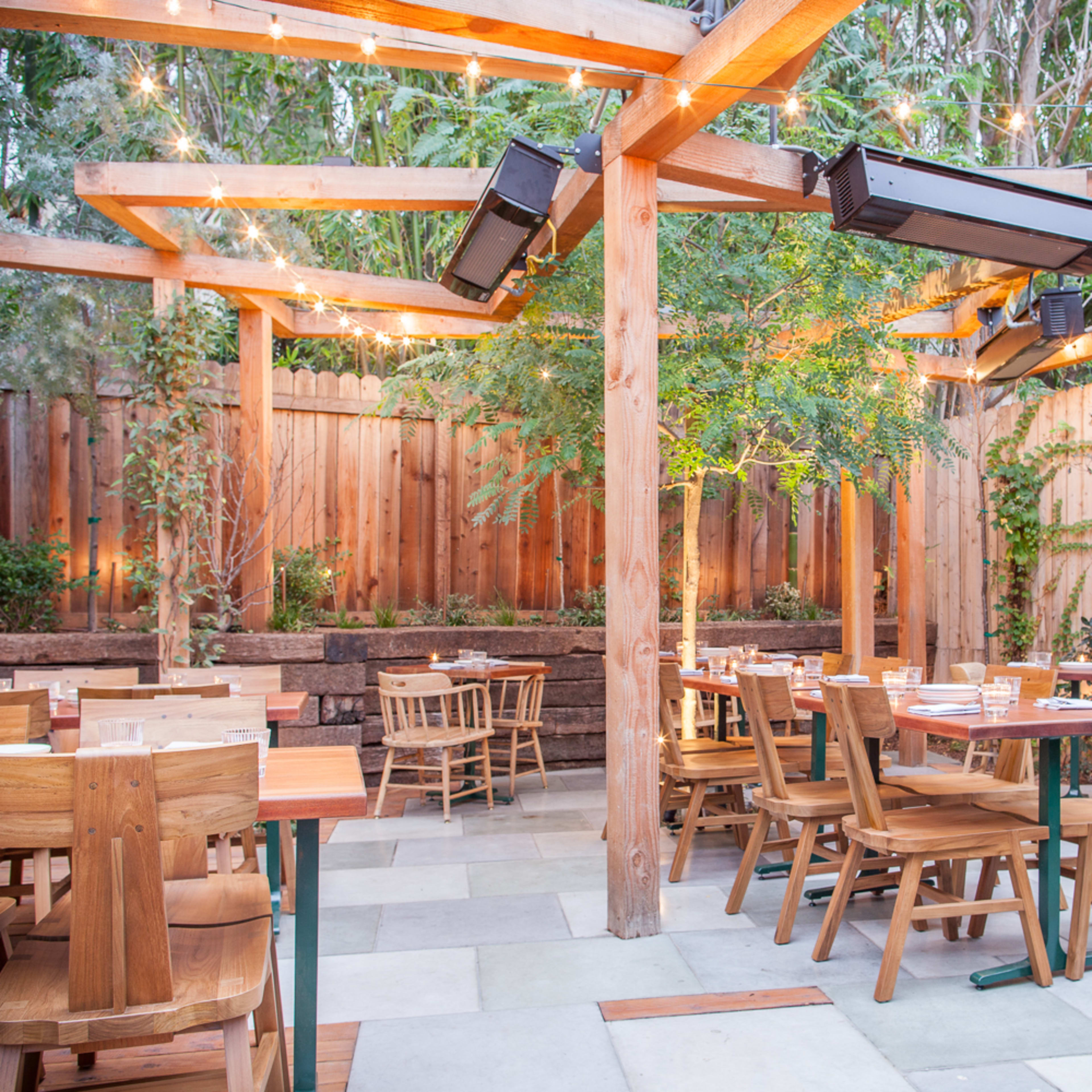An outdoor restaurant patio featuring wooden tables and chairs, surrounded by greenery and wooden fencing, with string lights overhead.