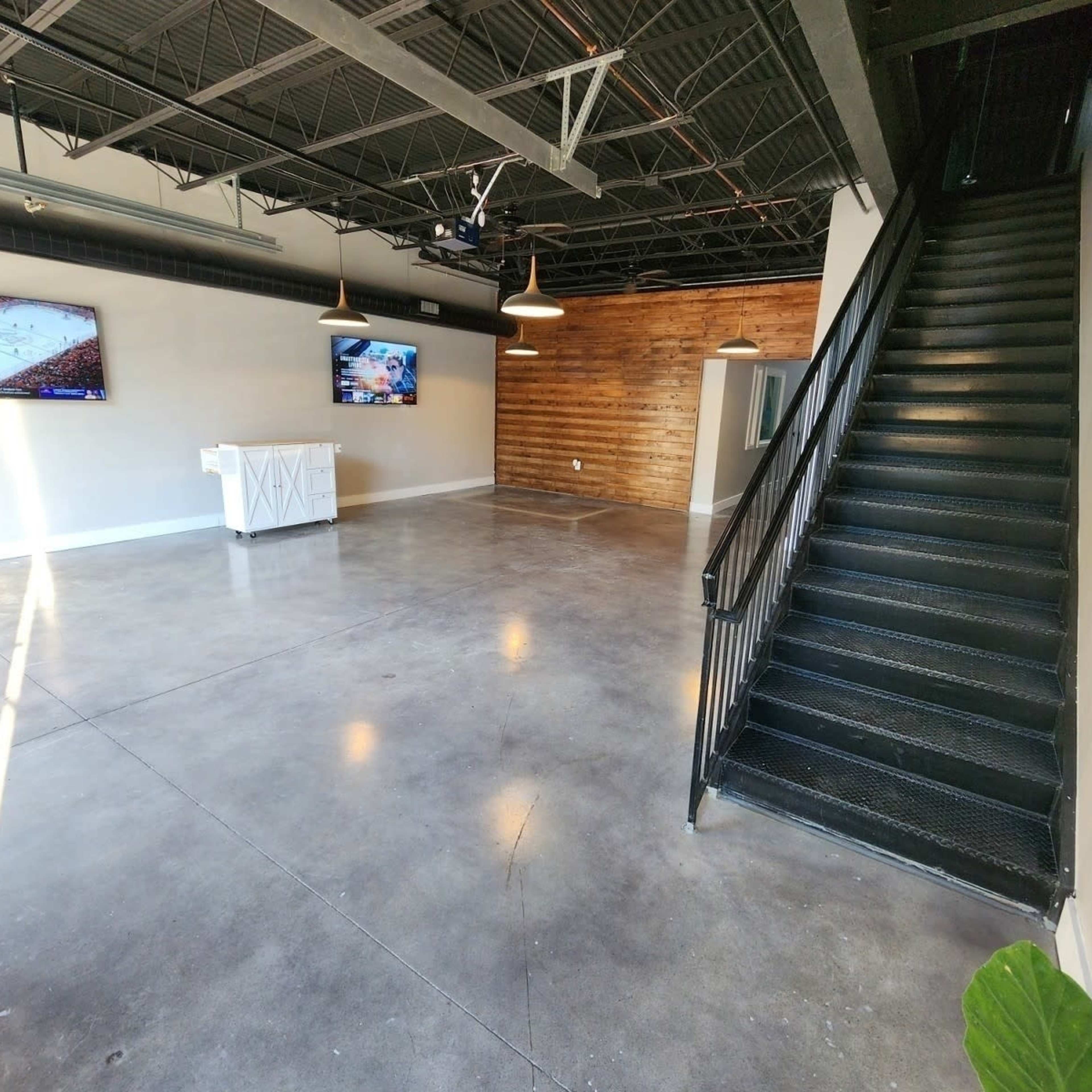 An empty room features a polished concrete floor, two wall-mounted televisions, a white reception-style desk, and a staircase leading to an upper level.