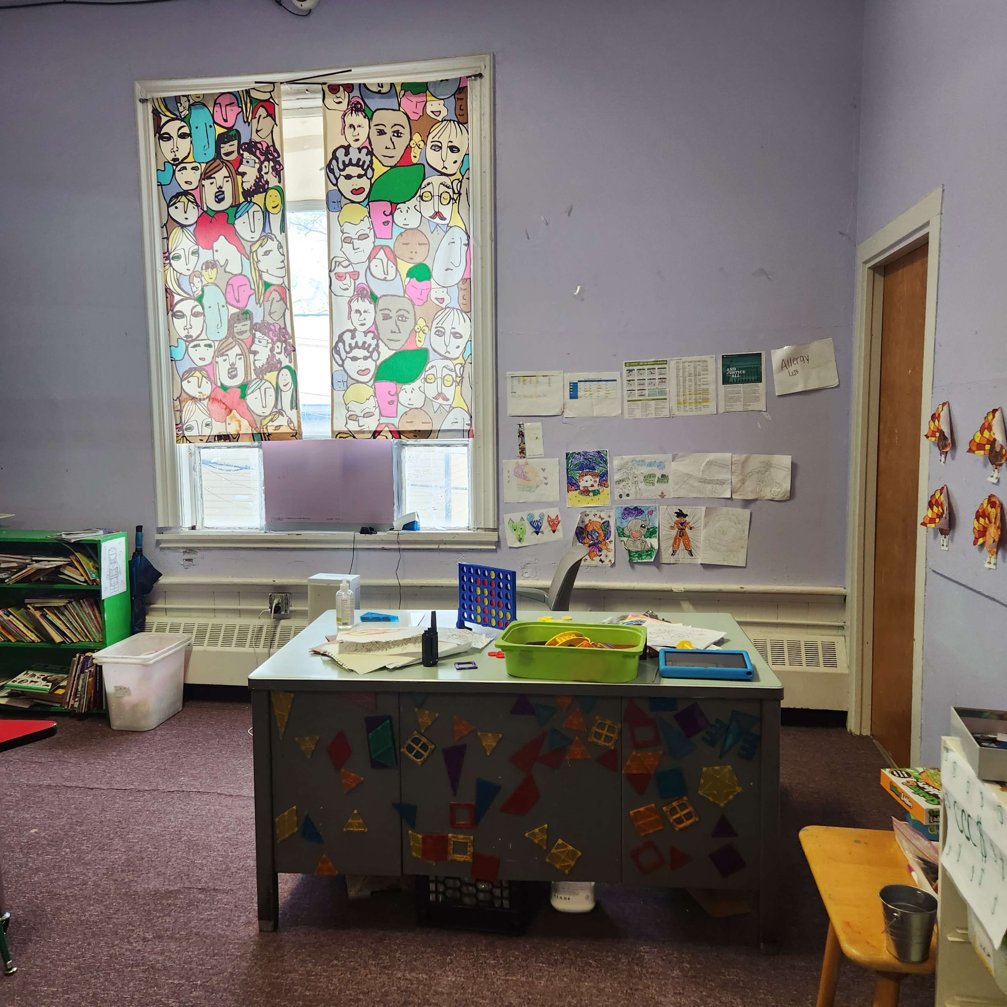 A classroom features a desk at the center, surrounded by bookshelves, colorful artwork on the walls, and patterned curtains.