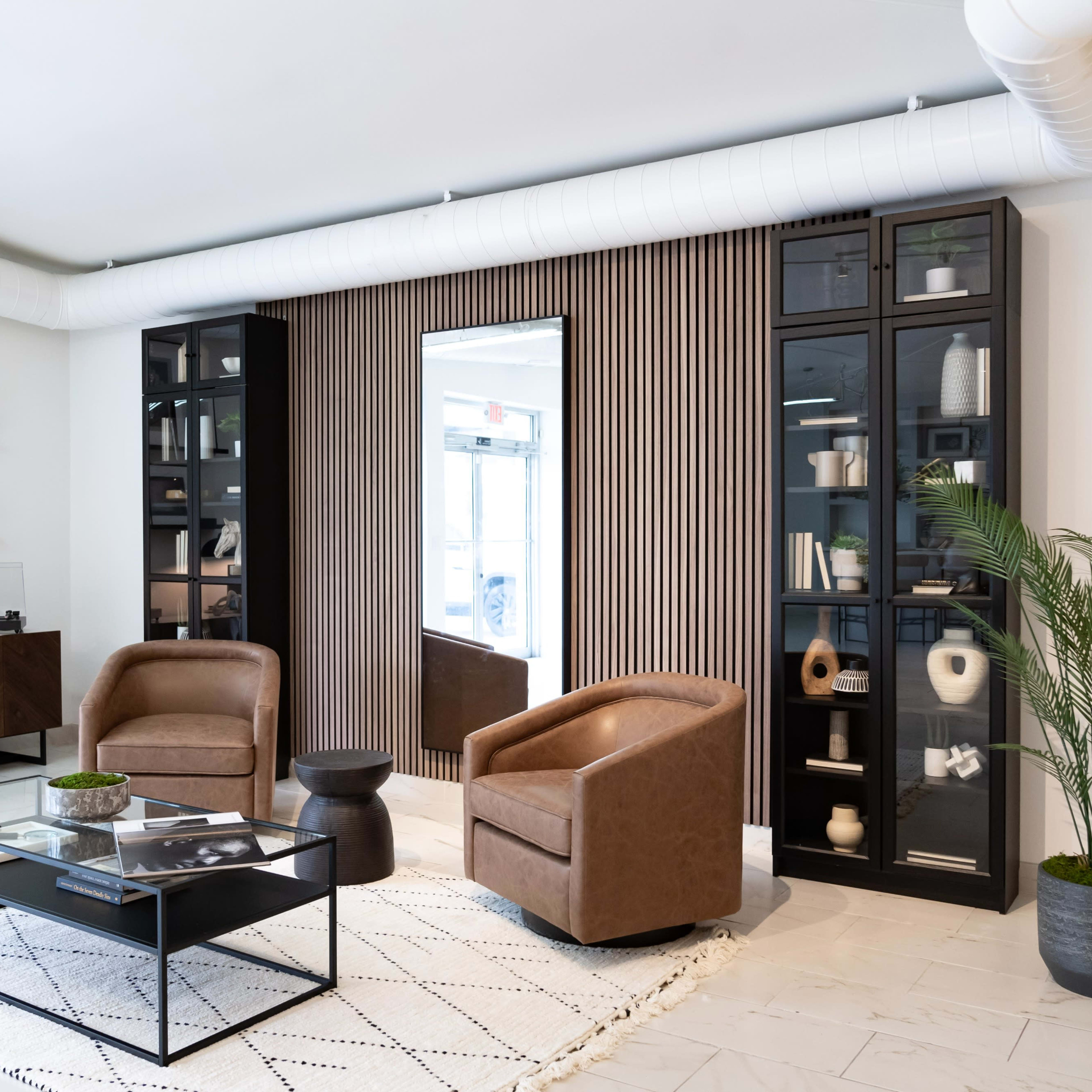 A modern living room set with two brown leather chairs, a glass coffee table, a patterned rug, and shelves displaying decorative items against a wall with vertical wood paneling.