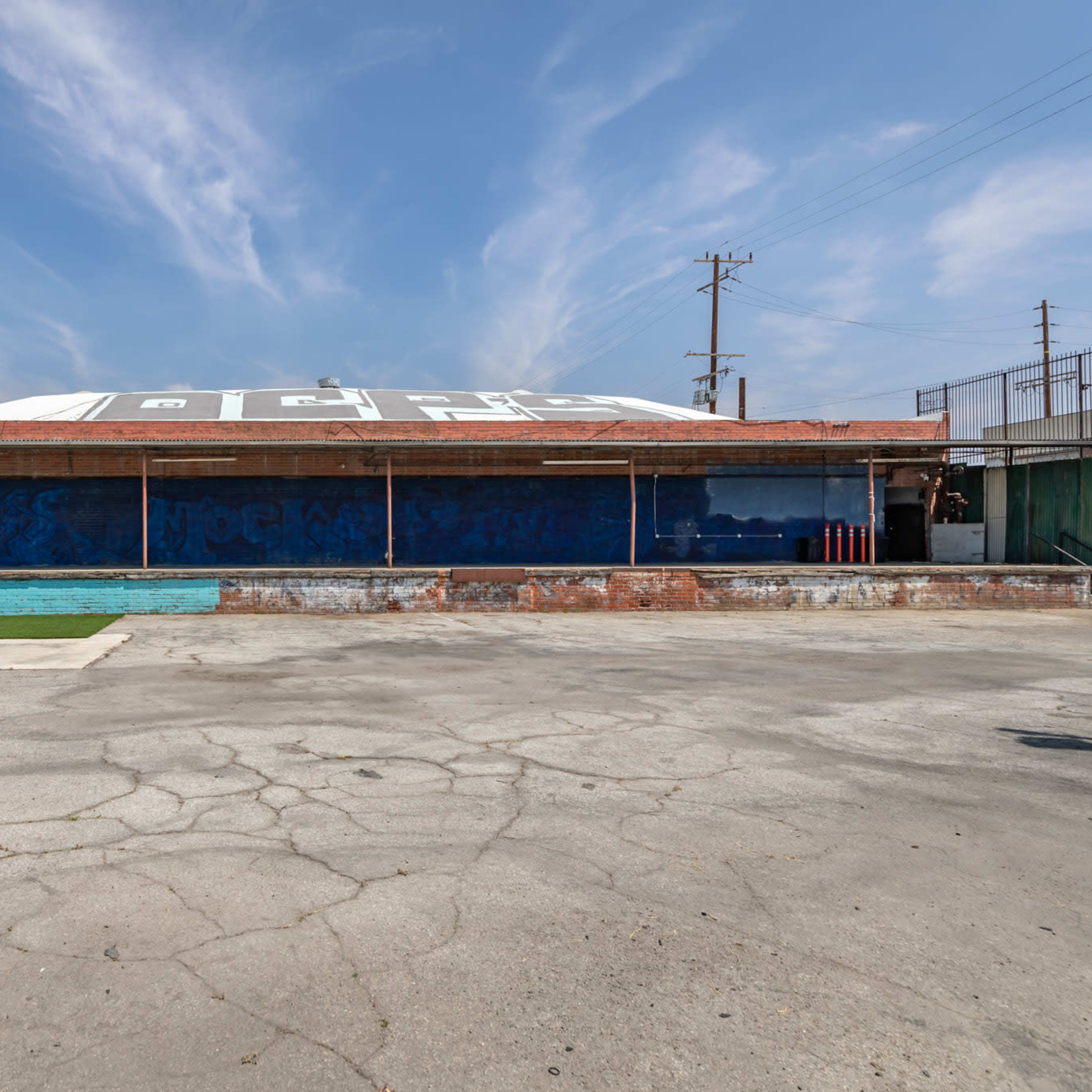 An empty outdoor area with a faded, mural-covered wall, a pool, and blue seating against a clear sky.