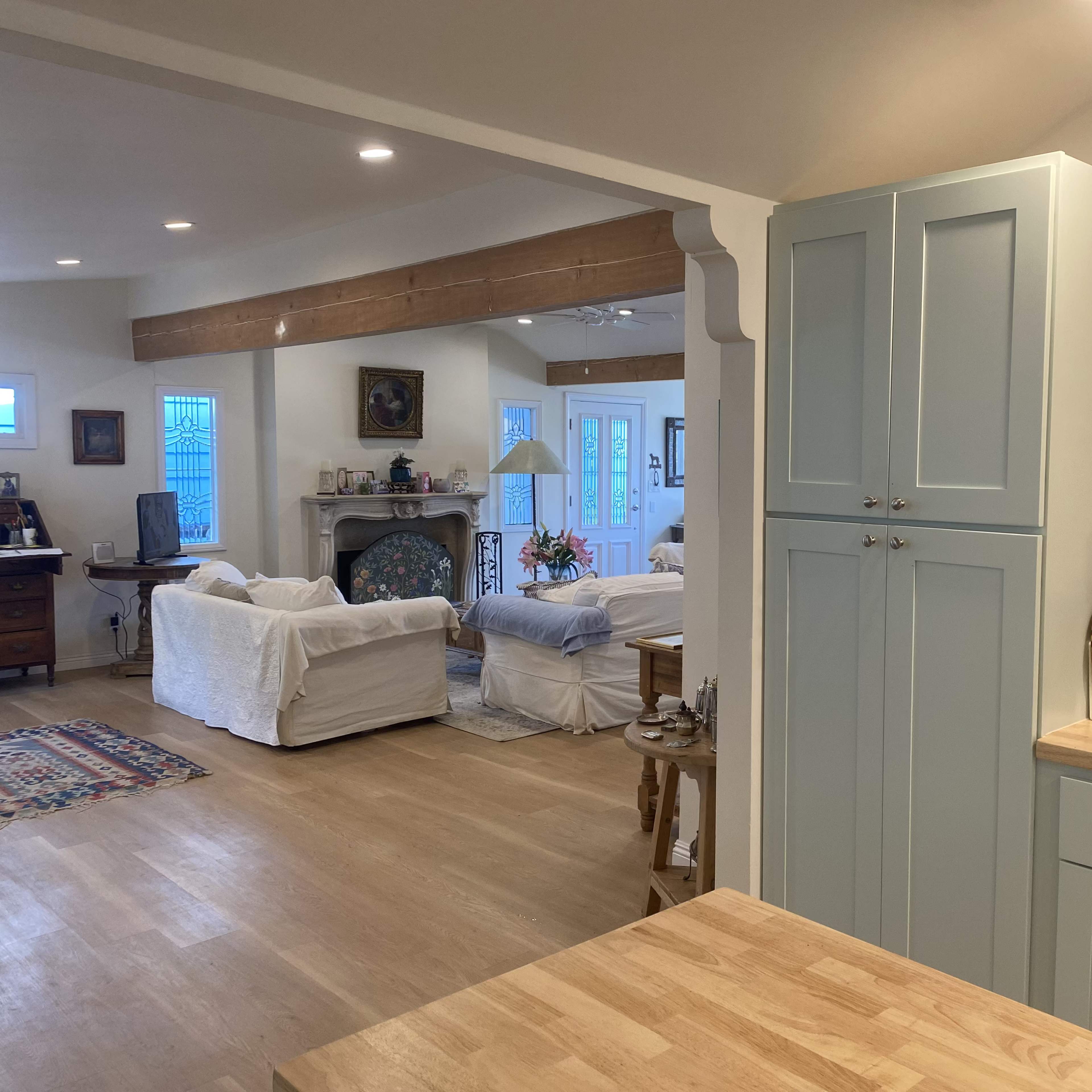 A modern kitchen with light blue cabinetry and a wooden countertop, opening to a living area furnished with white-covered sofas.