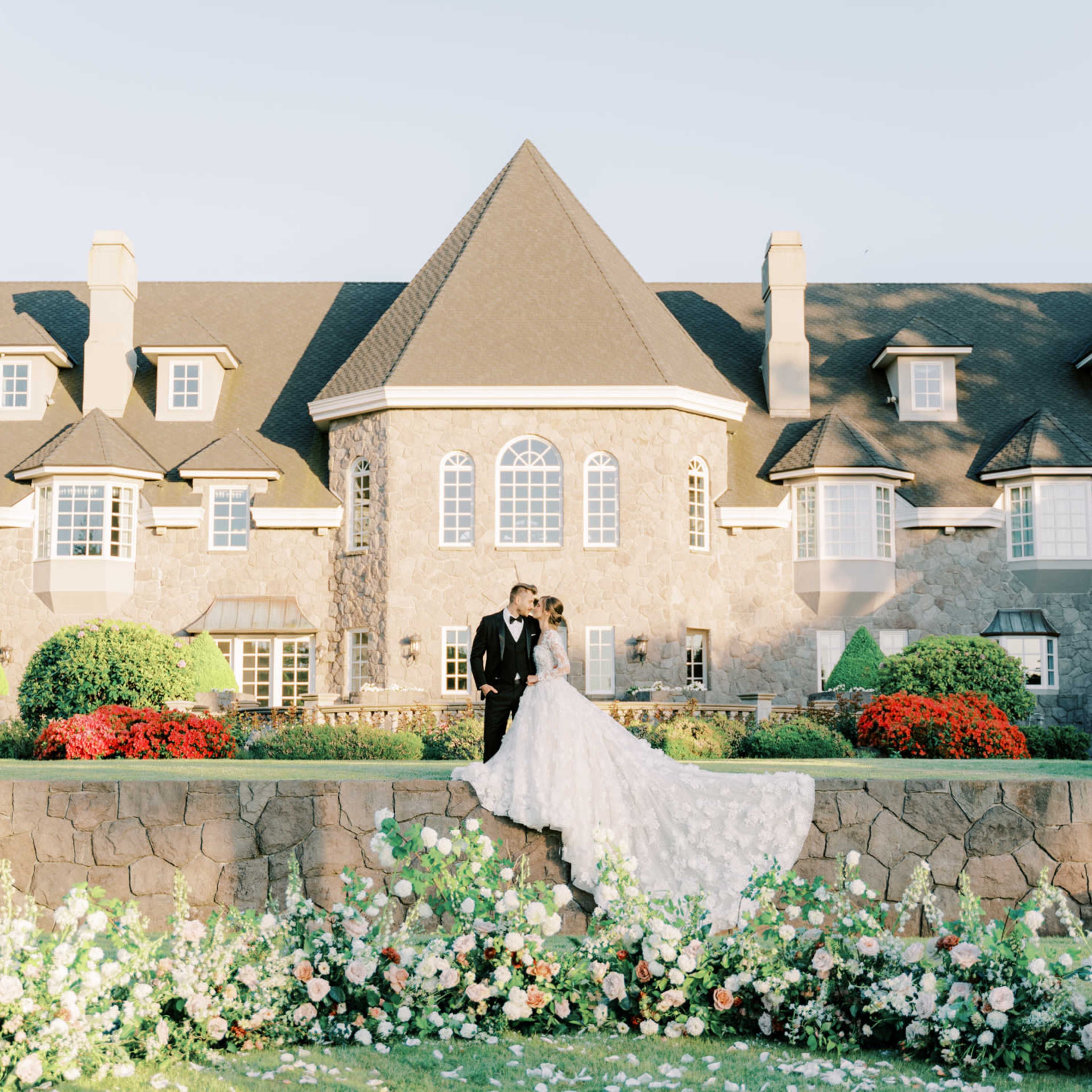 A couple in wedding attire stands together on a stone terrace in front of a large, intricately designed mansion surrounded by well-maintained flower beds.
