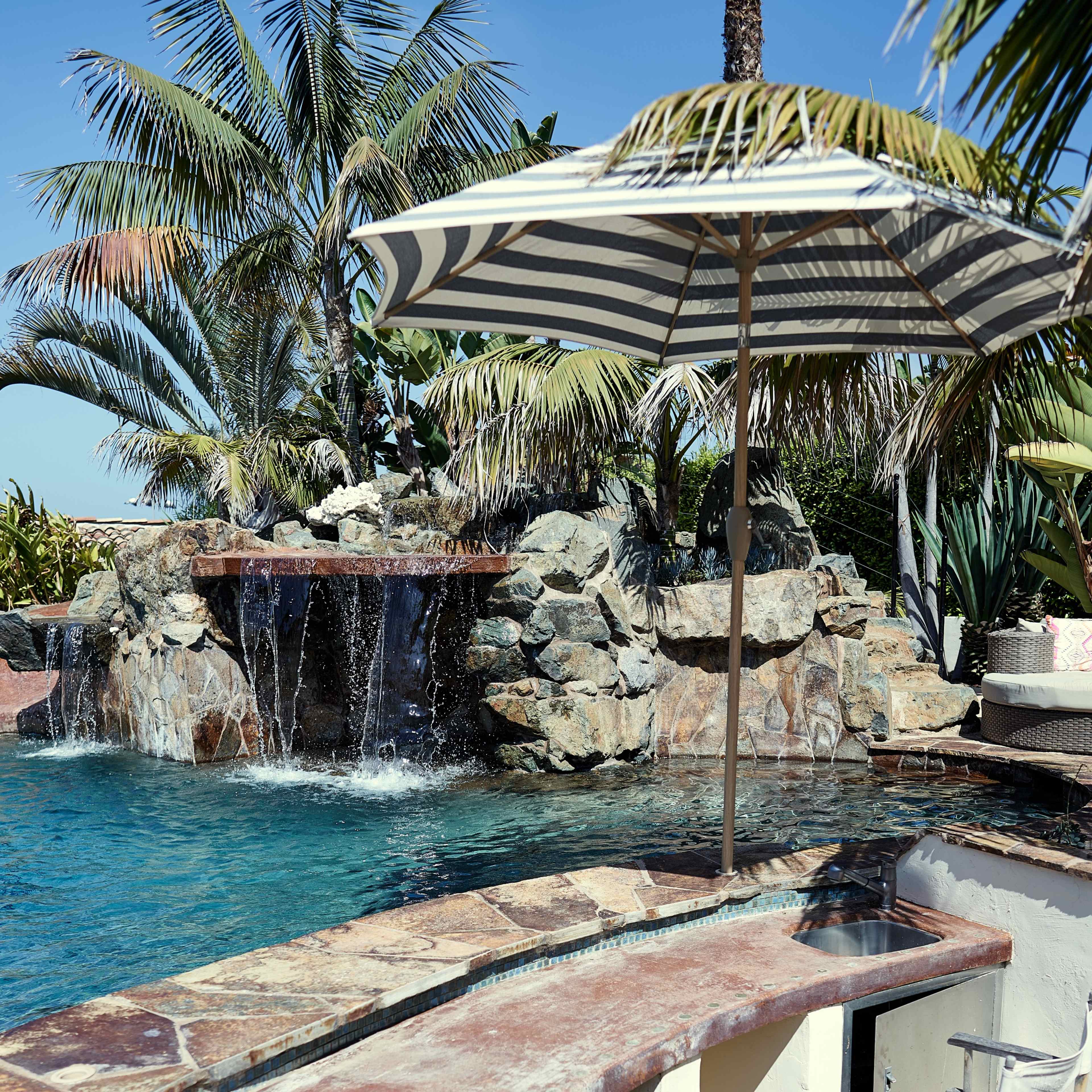 A swimming pool with a waterfall feature is surrounded by palm trees and includes a stone bar with two director chairs and an umbrella.