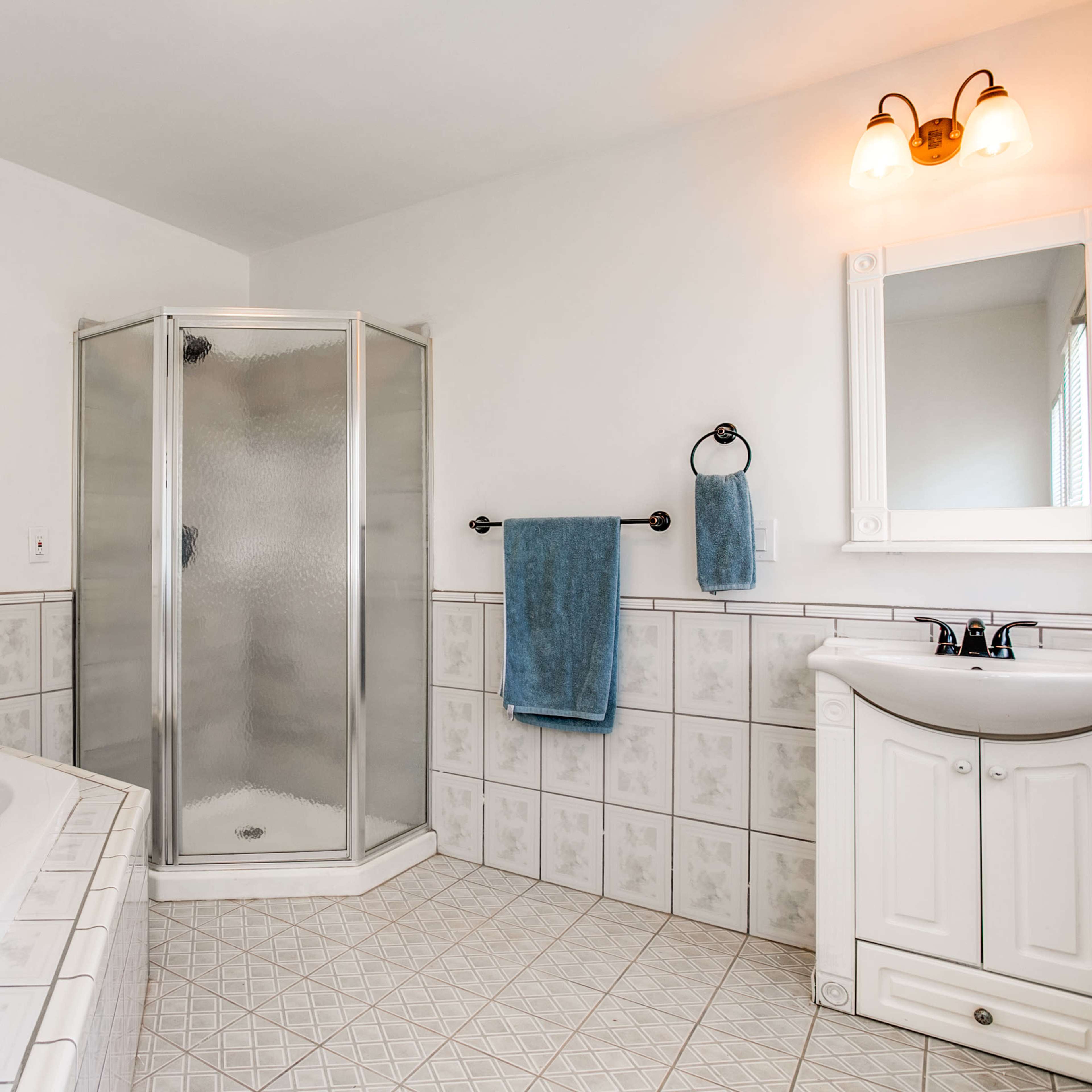 A bathroom with a corner shower, a large bathtub, and a sink with a cabinet, all surrounded by patterned tiles and a large window.