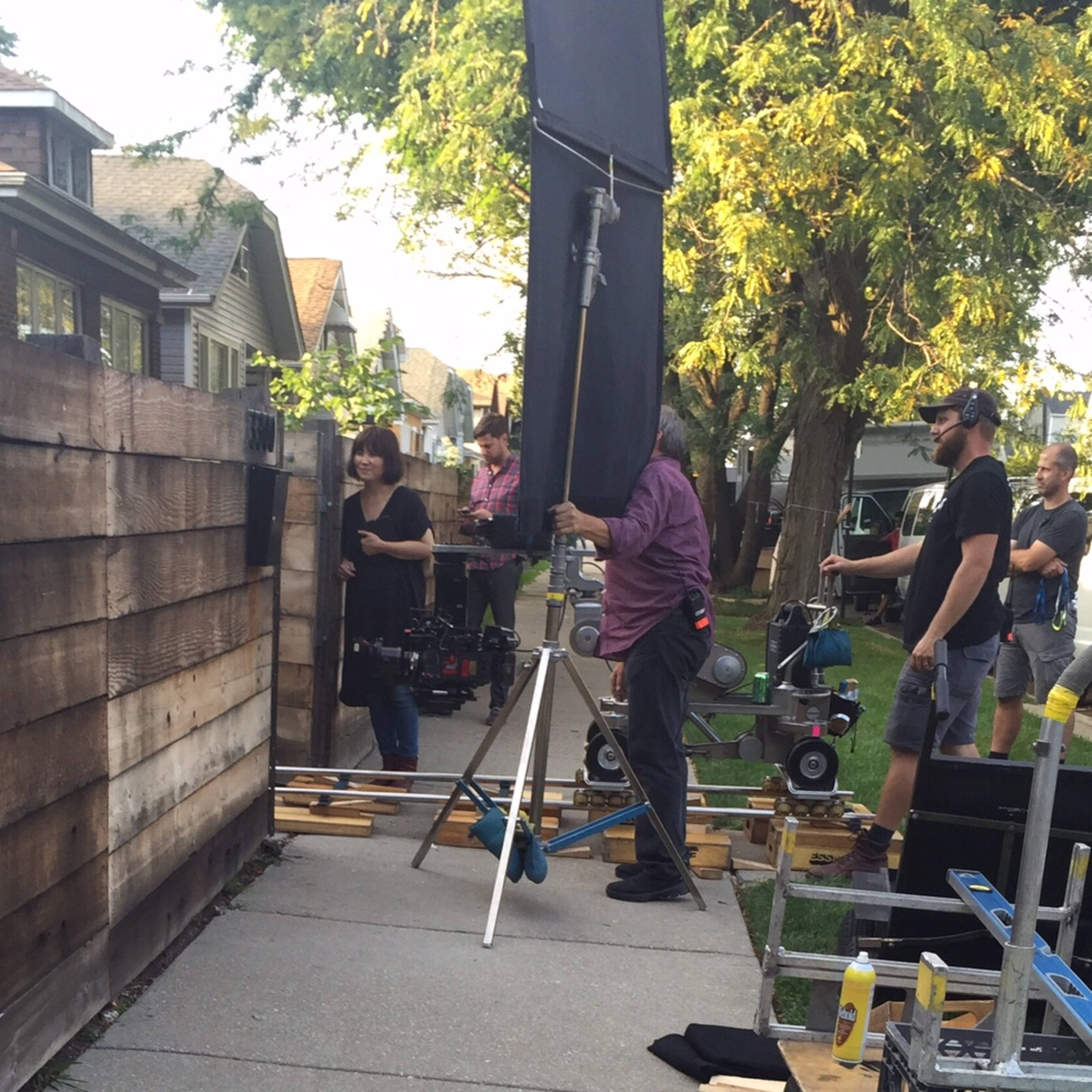A film crew is set up on a sidewalk near a wooden fence, with equipment including a camera on tracks and lighting apparatus.