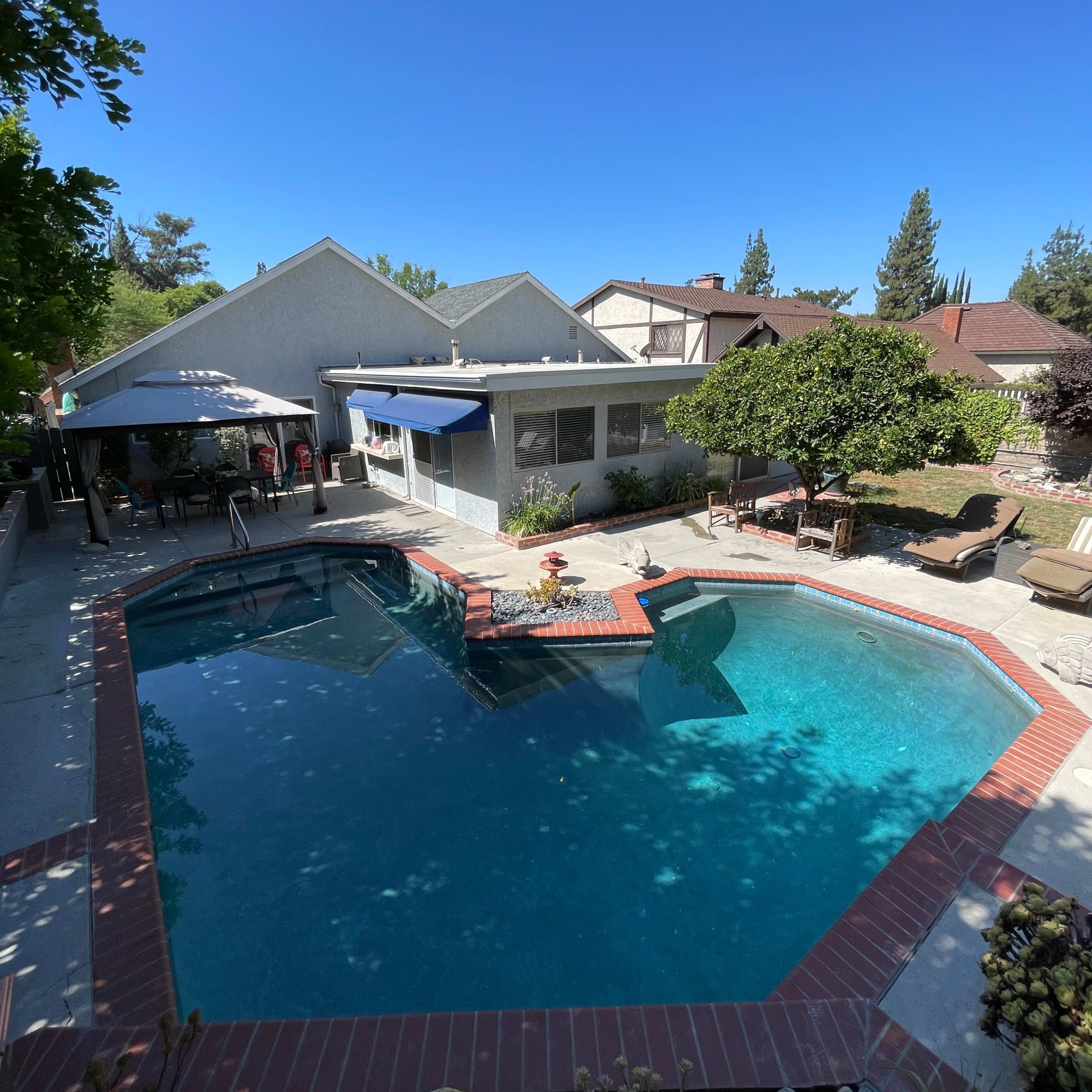 A backyard with a swimming pool surrounded by a patio area, lounge chairs, and a shaded seating space next to a house.