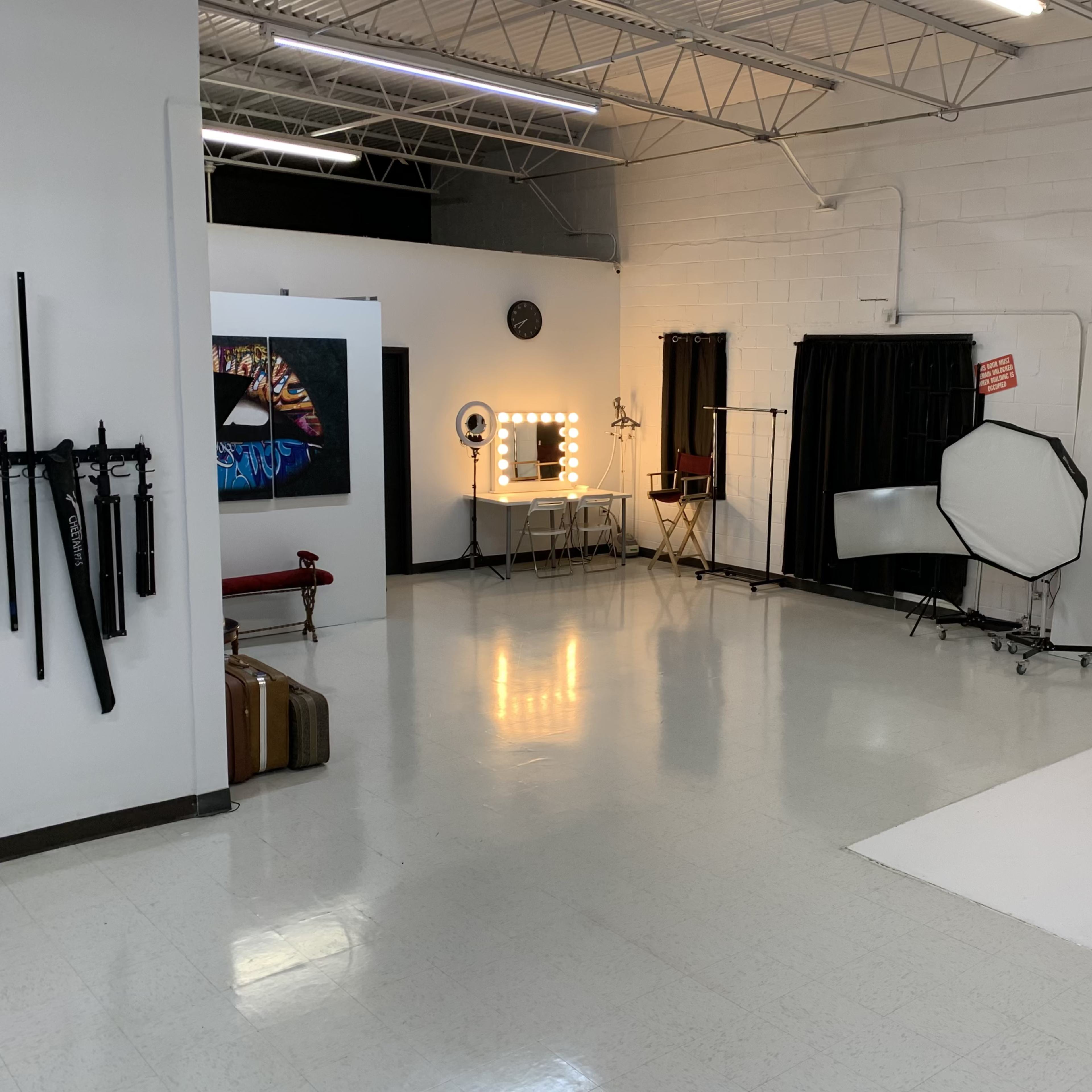 A photography studio with white walls, a large backdrop, a makeup station with lights, and various photography equipment.