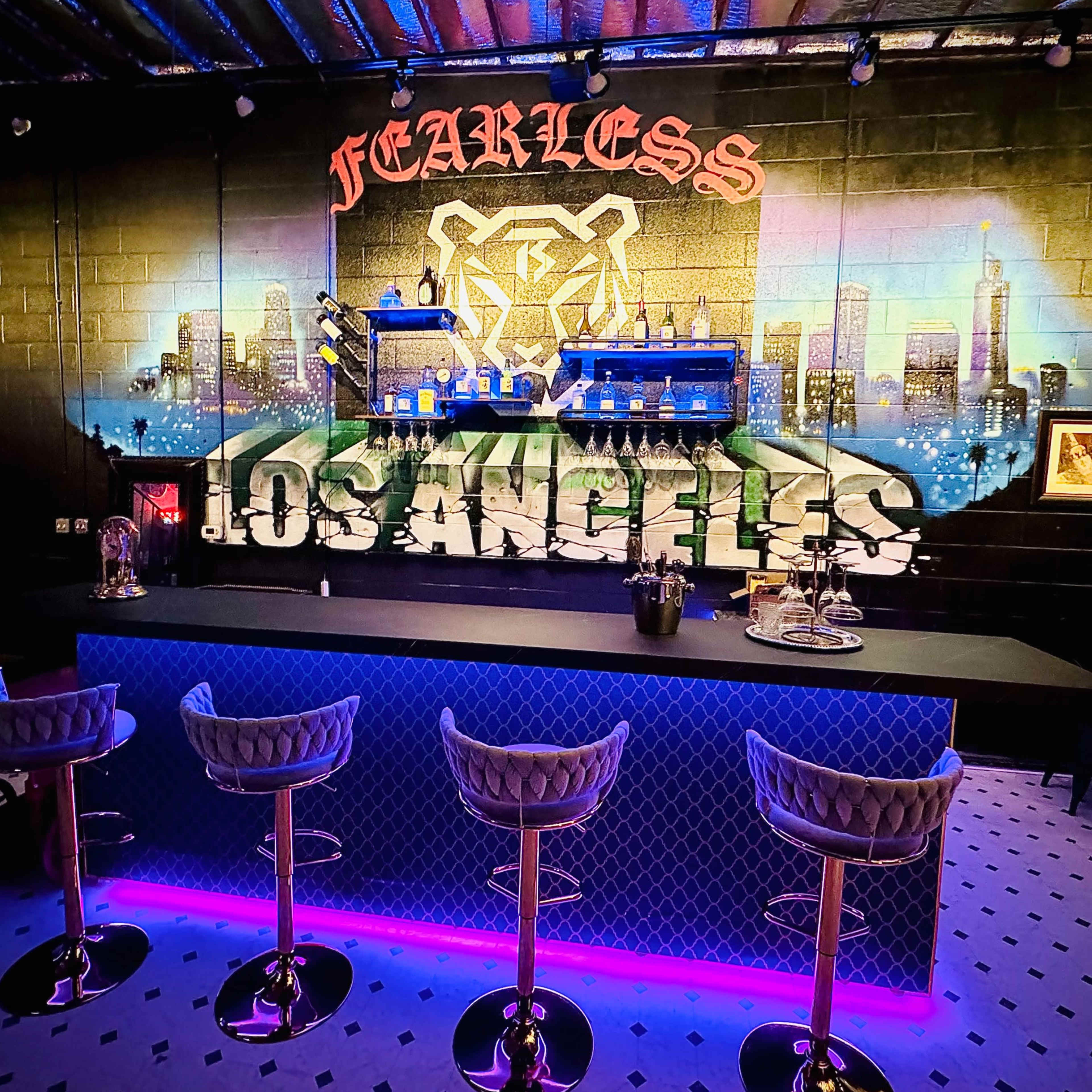 A modern bar area with a mural featuring a bear and "LOS ANGELES," illuminated by blue and purple lighting against a black wall.