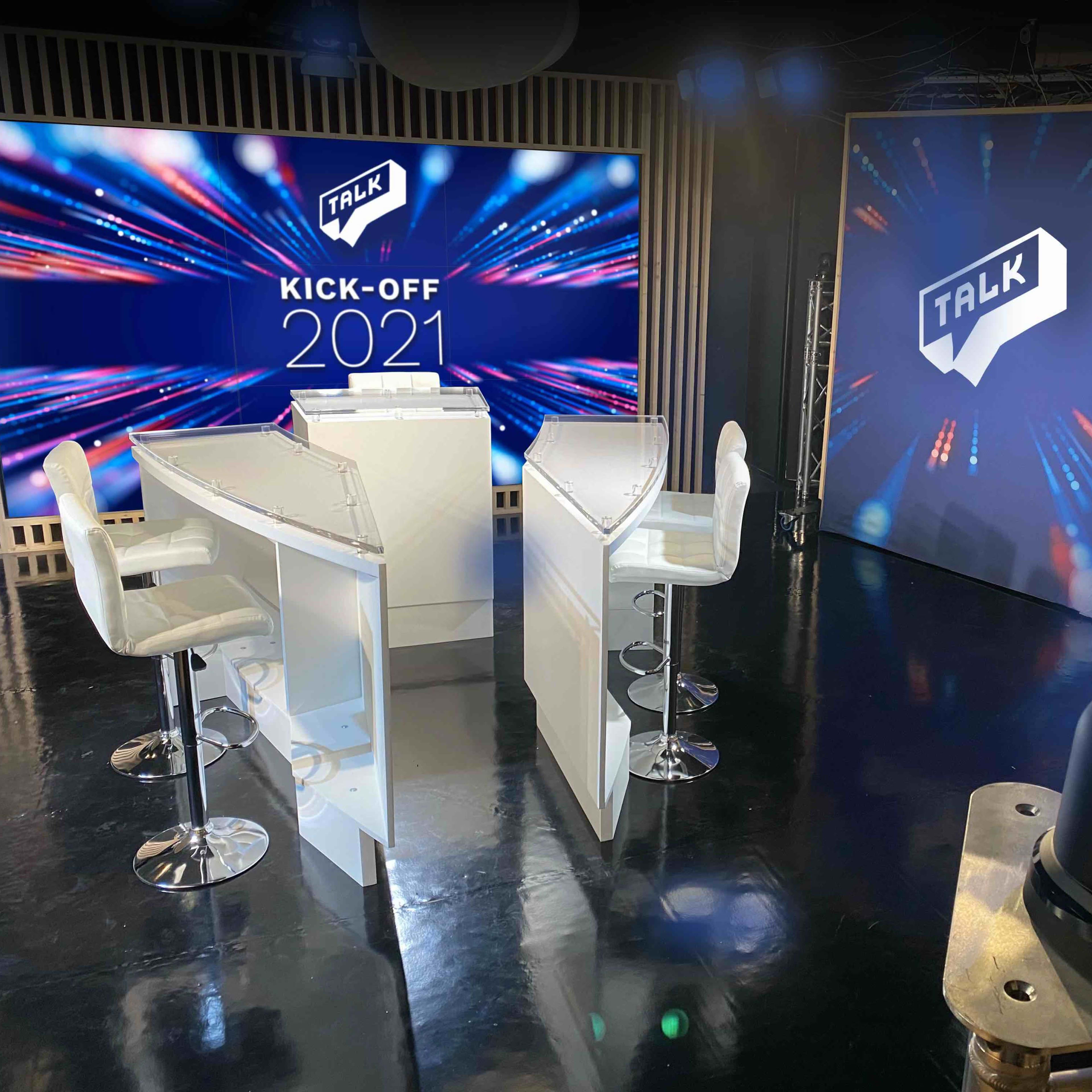 A modern broadcast studio setup with two white discussion tables, white chairs, and large screens displaying colorful graphics, labeled "KICK-OFF 2021" and "TALK."