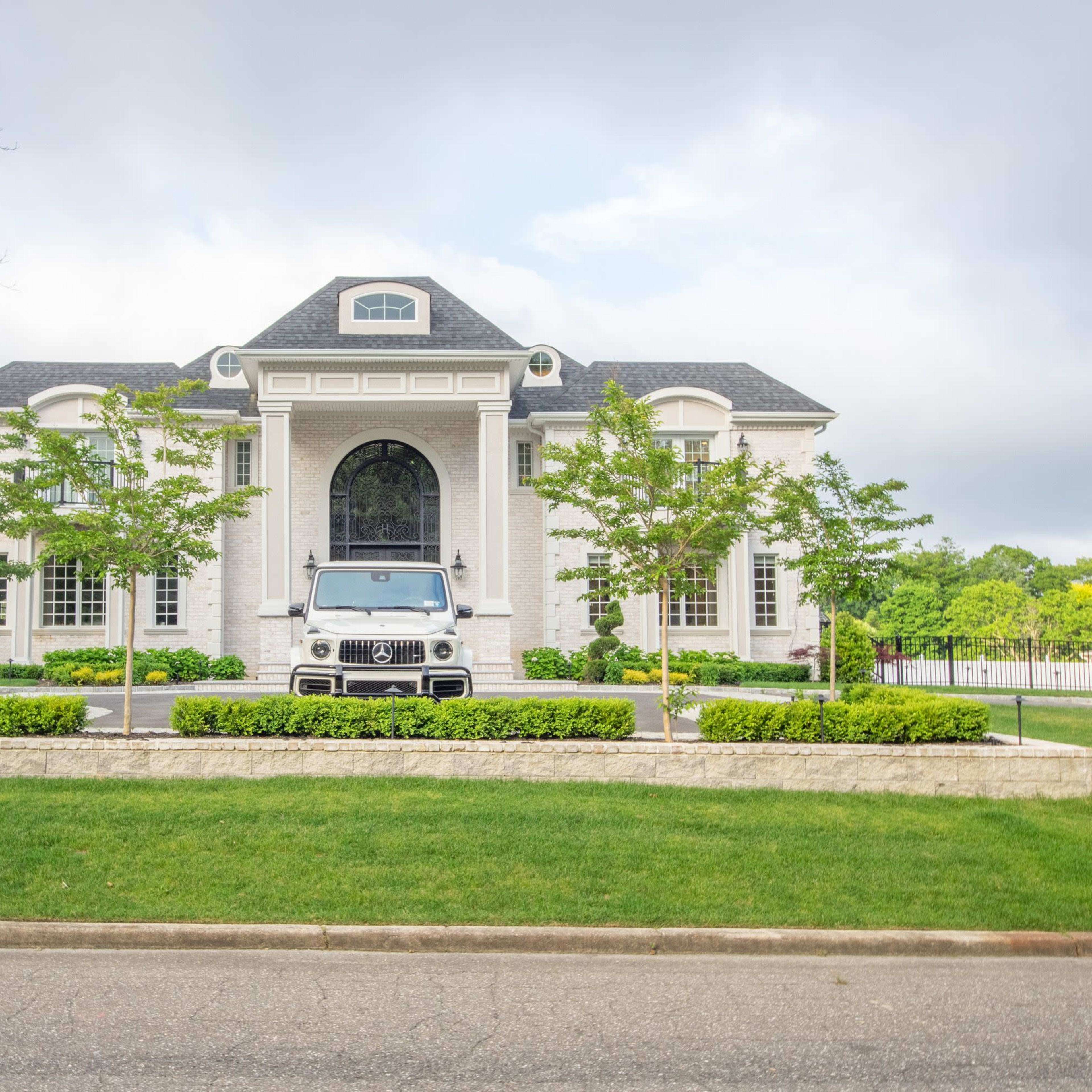 A large, white brick mansion with a front driveway and a parked vehicle is framed by well-maintained landscaping.