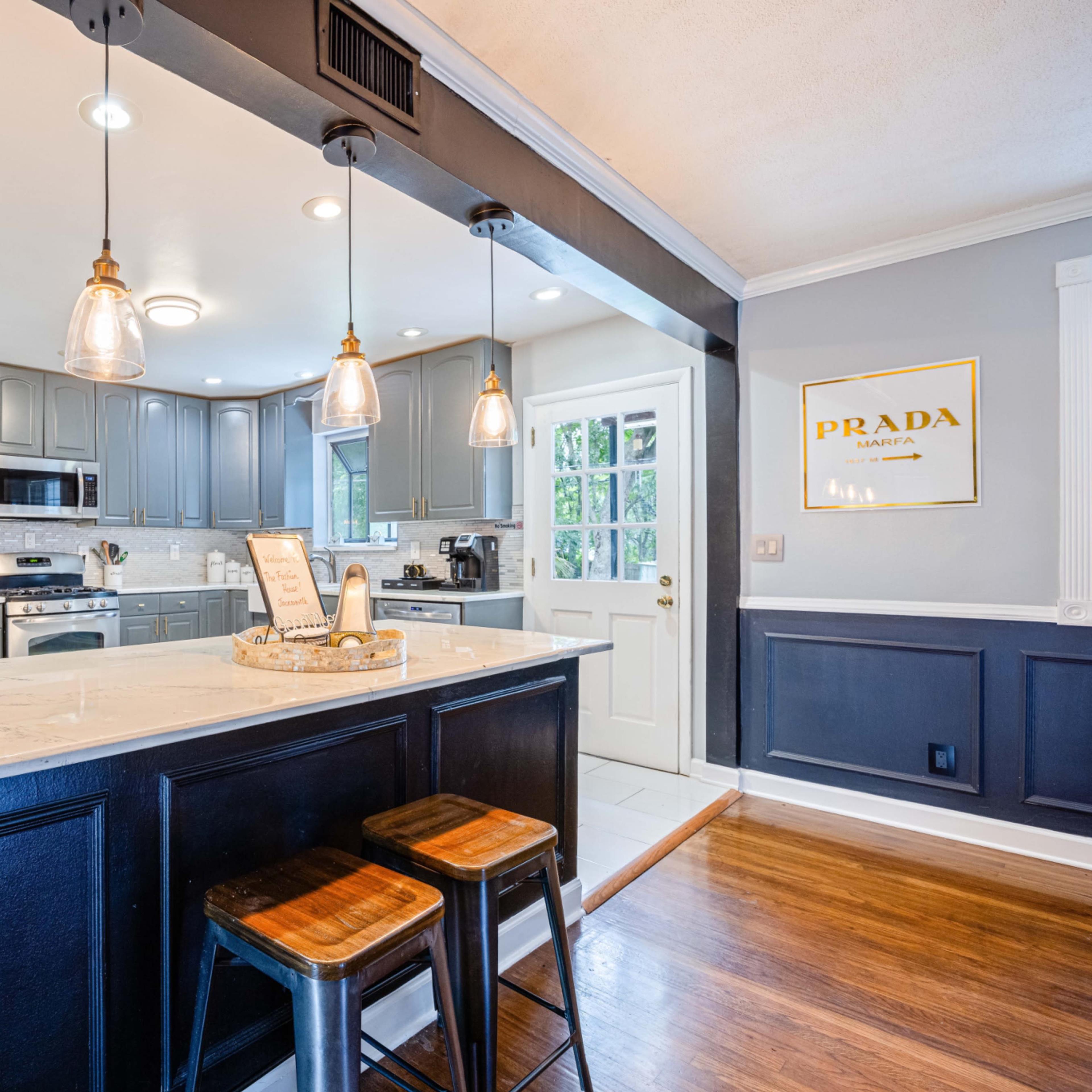 A modern kitchen features gray cabinets, stainless steel appliances, and a bar area with three wooden stools illuminated by pendant lights.