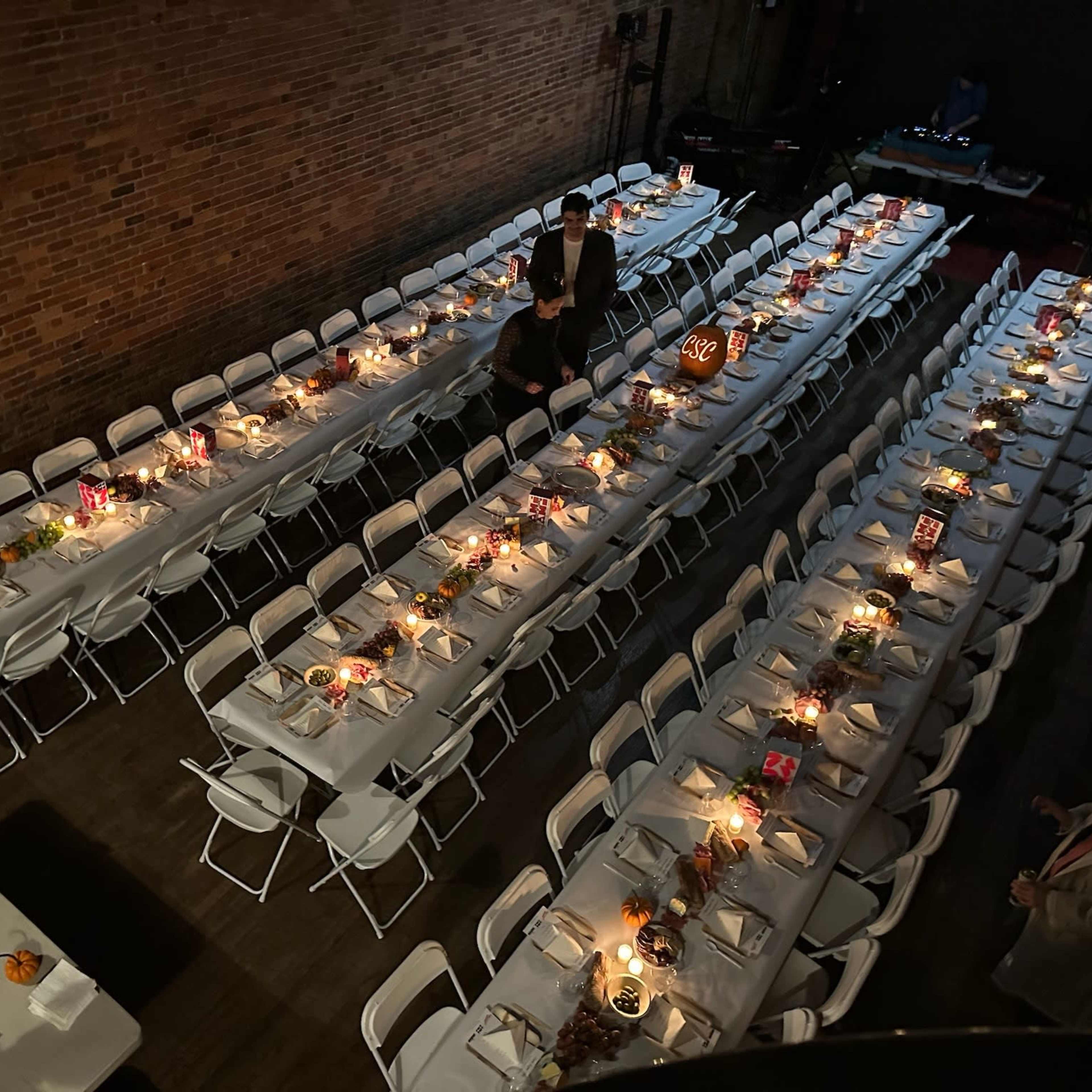 A large dining setup with multiple long tables arranged in rows, each decorated with candles and centerpieces, in a space with exposed brick walls.