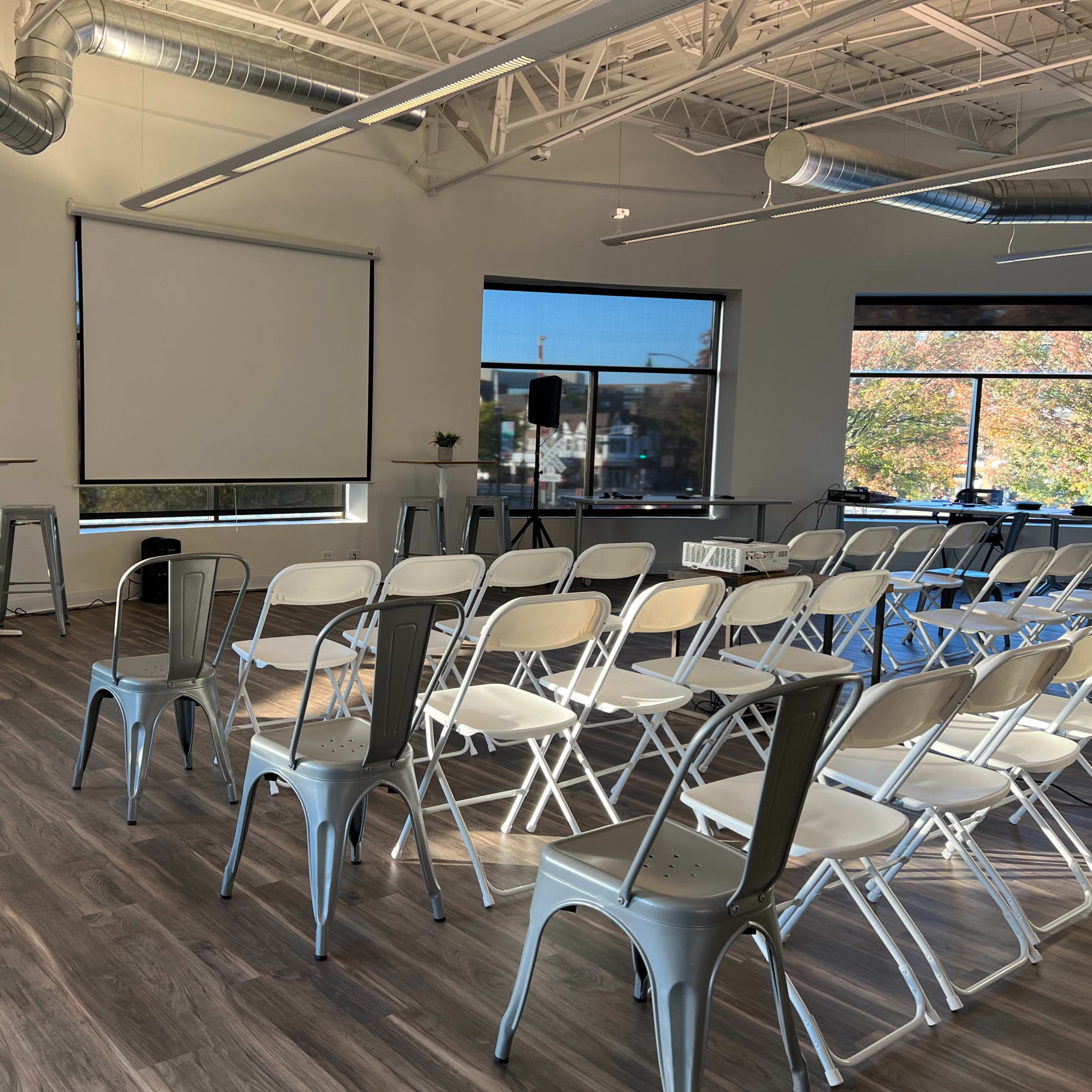 A conference room set up with rows of chairs facing a blank presentation screen, alongside a table on a stage and large windows revealing an outdoor view.