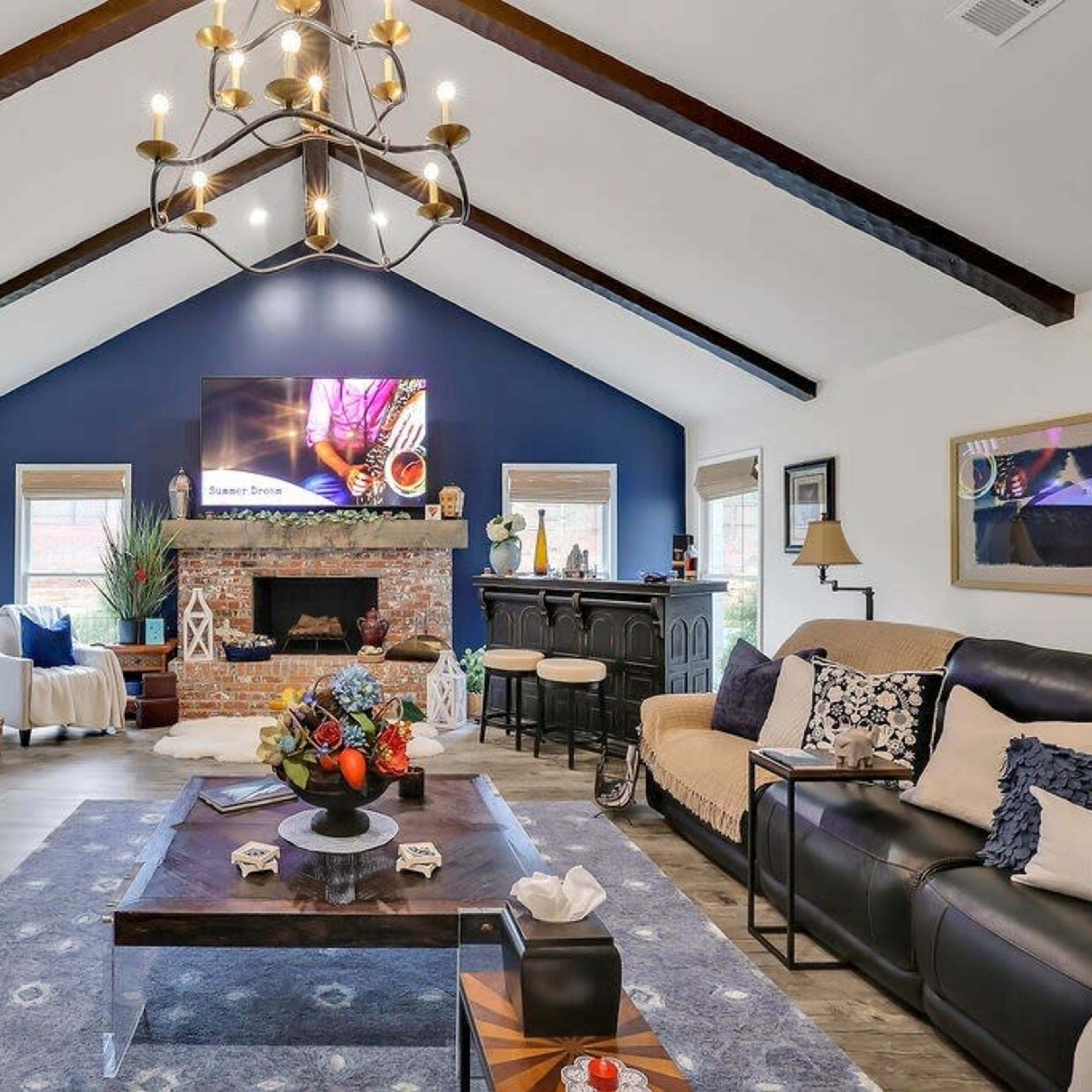 The living room features a vaulted ceiling with exposed beams, a large flat-screen TV mounted on a dark blue wall, and a central coffee table surrounded by sofas and decorative accents.