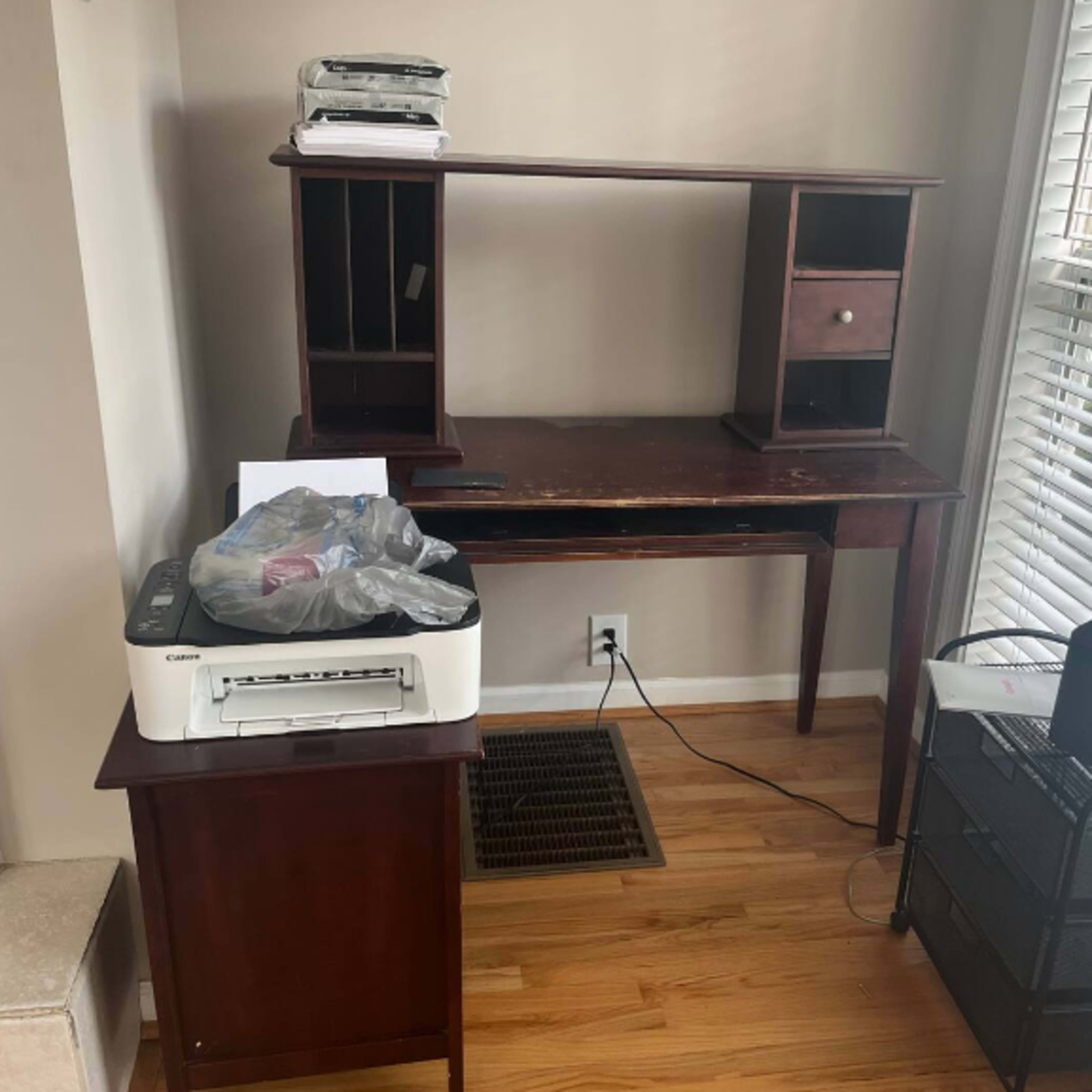 A wooden desk with a shelf unit, a printer, and a small storage cart is positioned near a window with blinds.