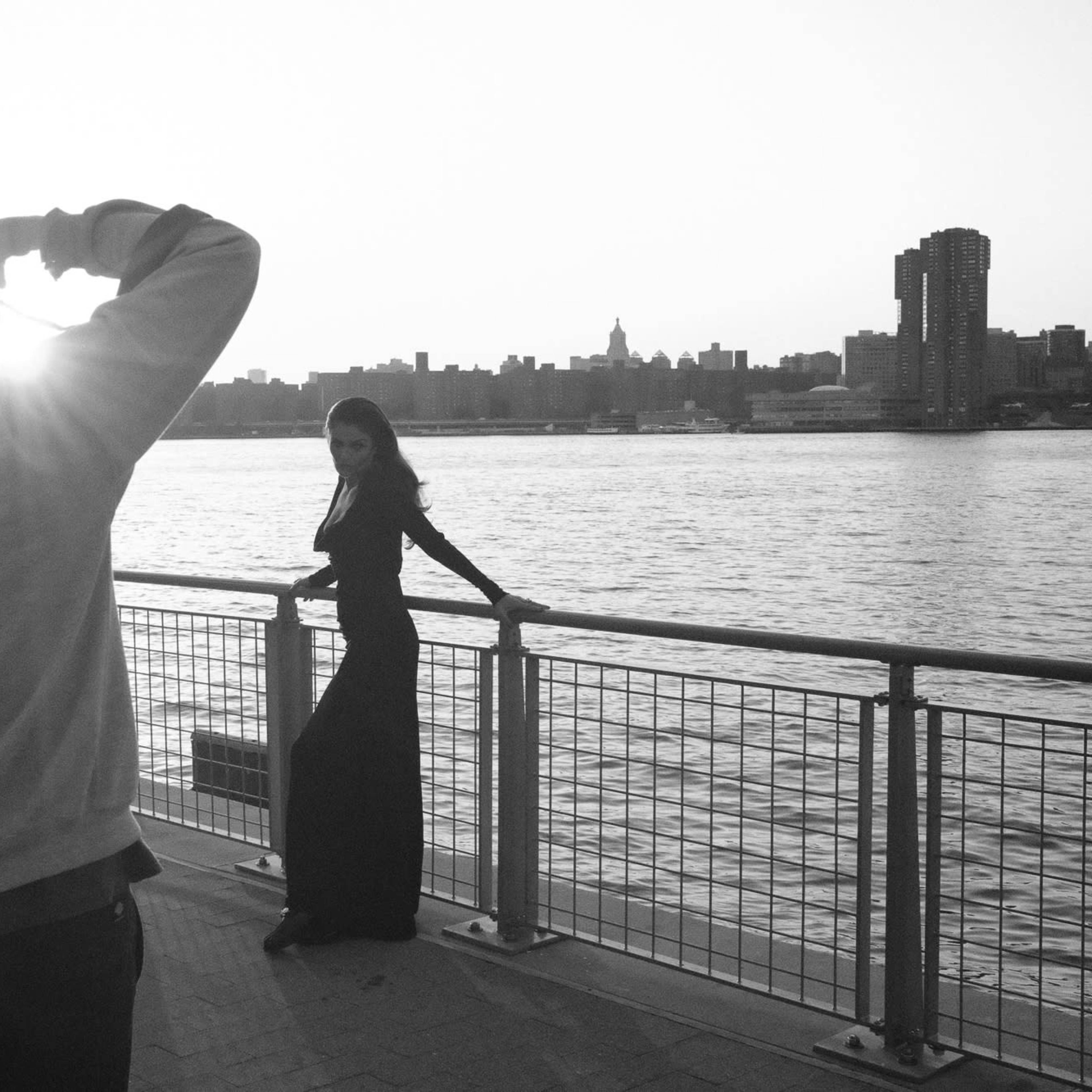 A person takes a photograph of a woman standing by a waterfront with a city skyline in the background at sunset.