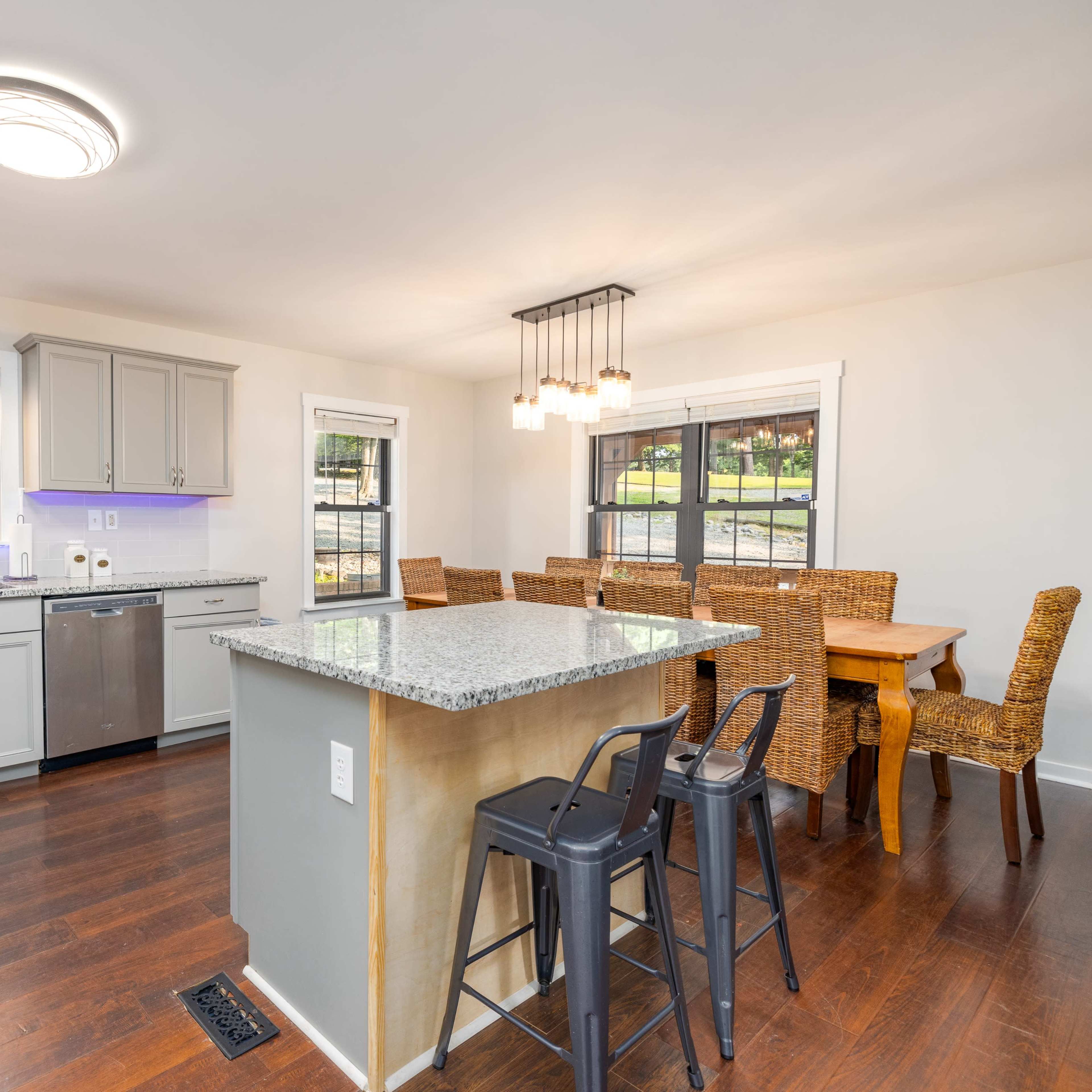 A modern kitchen features gray cabinets, a granite island, and a dining area with woven chairs and a wooden table.