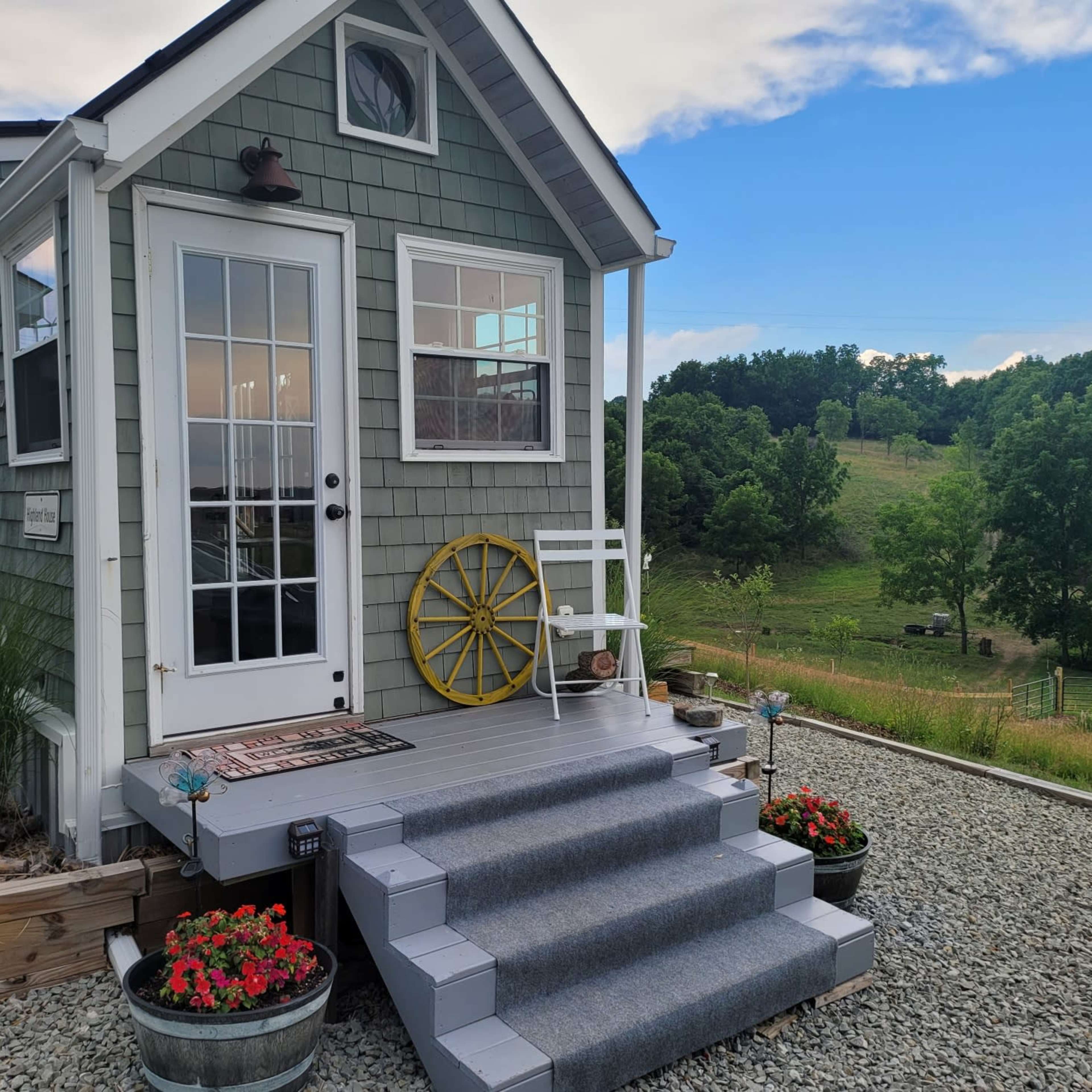 A small, green cottage with a front porch, a yellow wheel decoration, potted flowers, and a scenic view of trees and hills in the background.