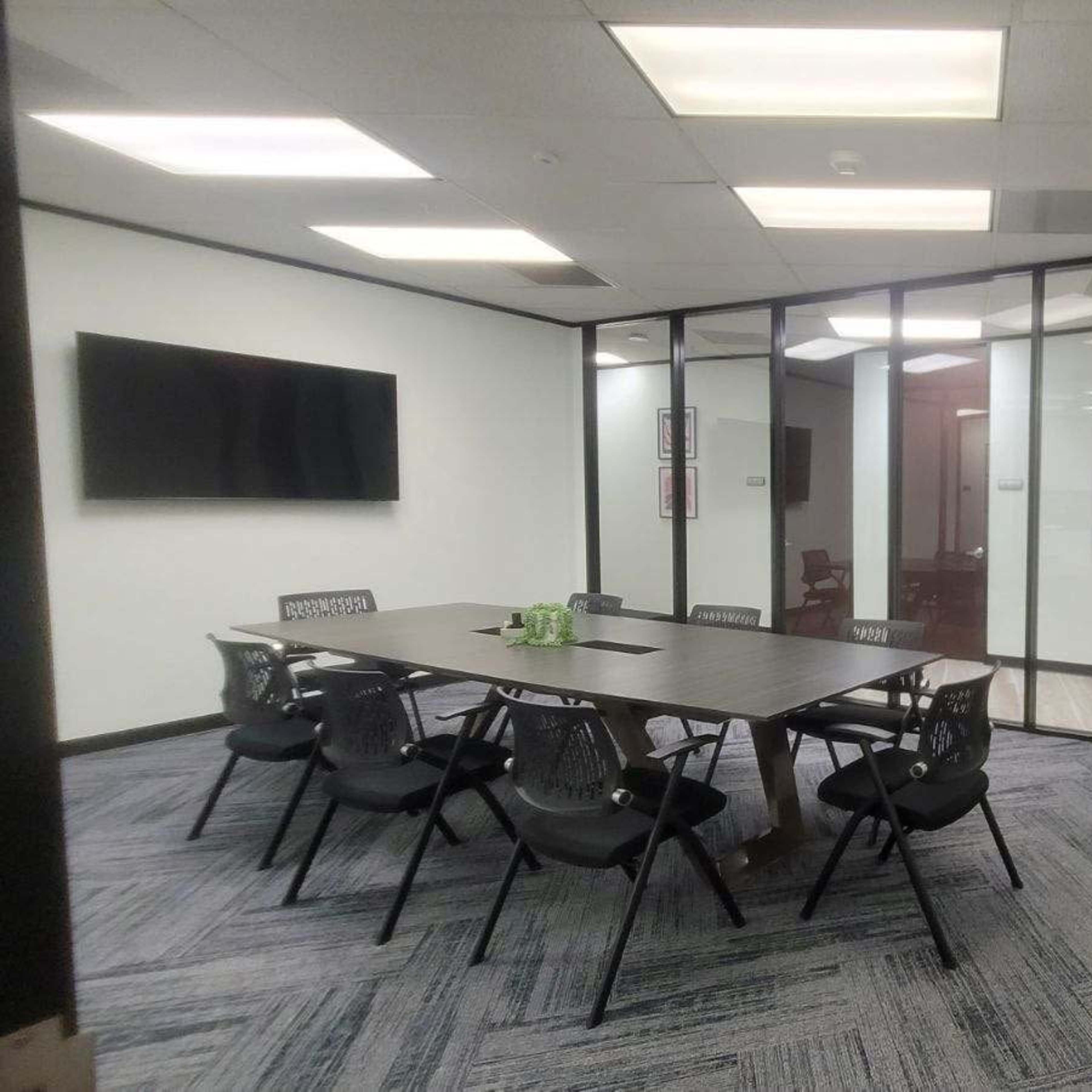 A modern conference room featuring a long table surrounded by black chairs, with glass walls and a television mounted on one side.