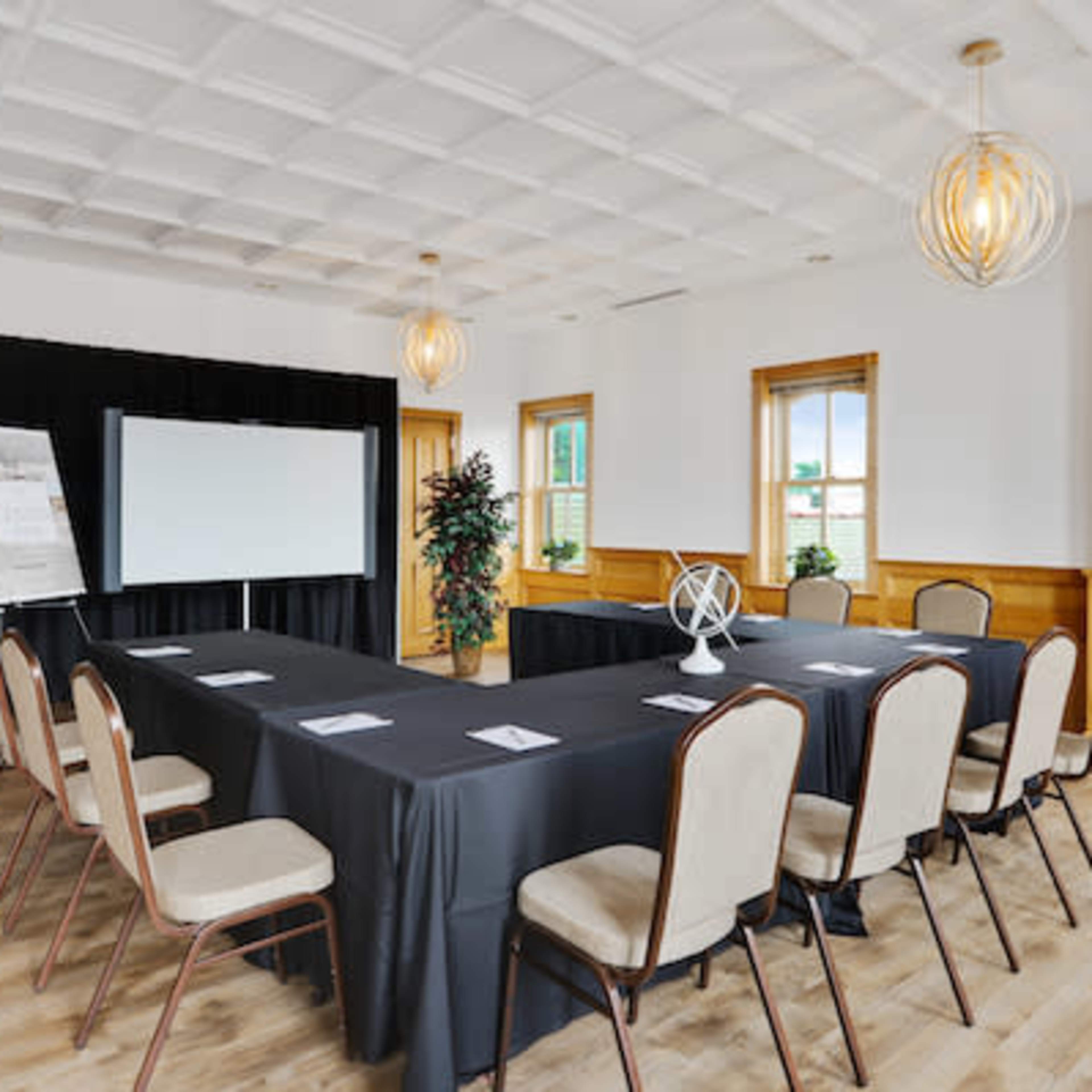 A conference room features a long black table surrounded by beige chairs, with a large screen at one end and windows allowing natural light.