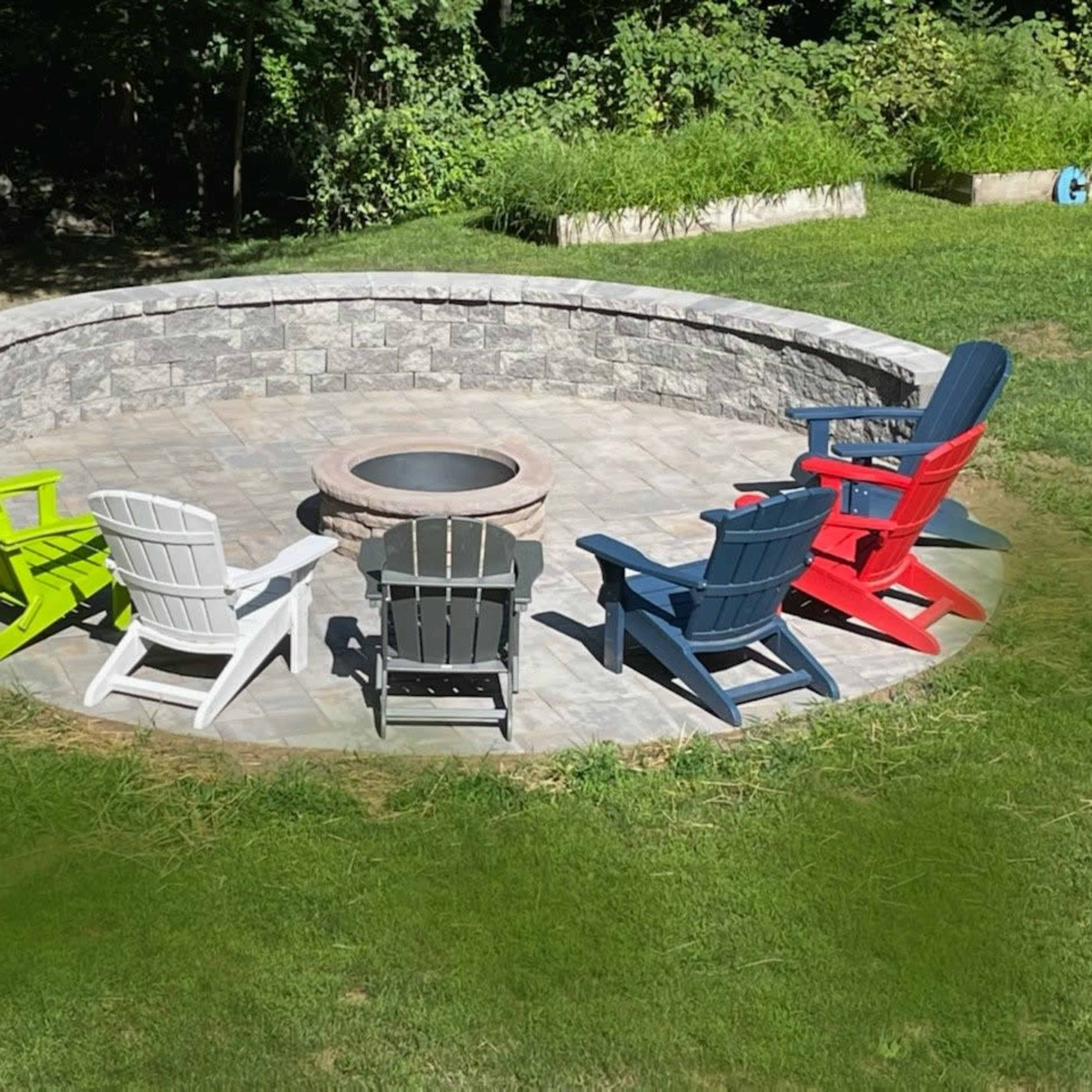 A circular stone patio features five brightly colored Adirondack chairs arranged around a fire pit.