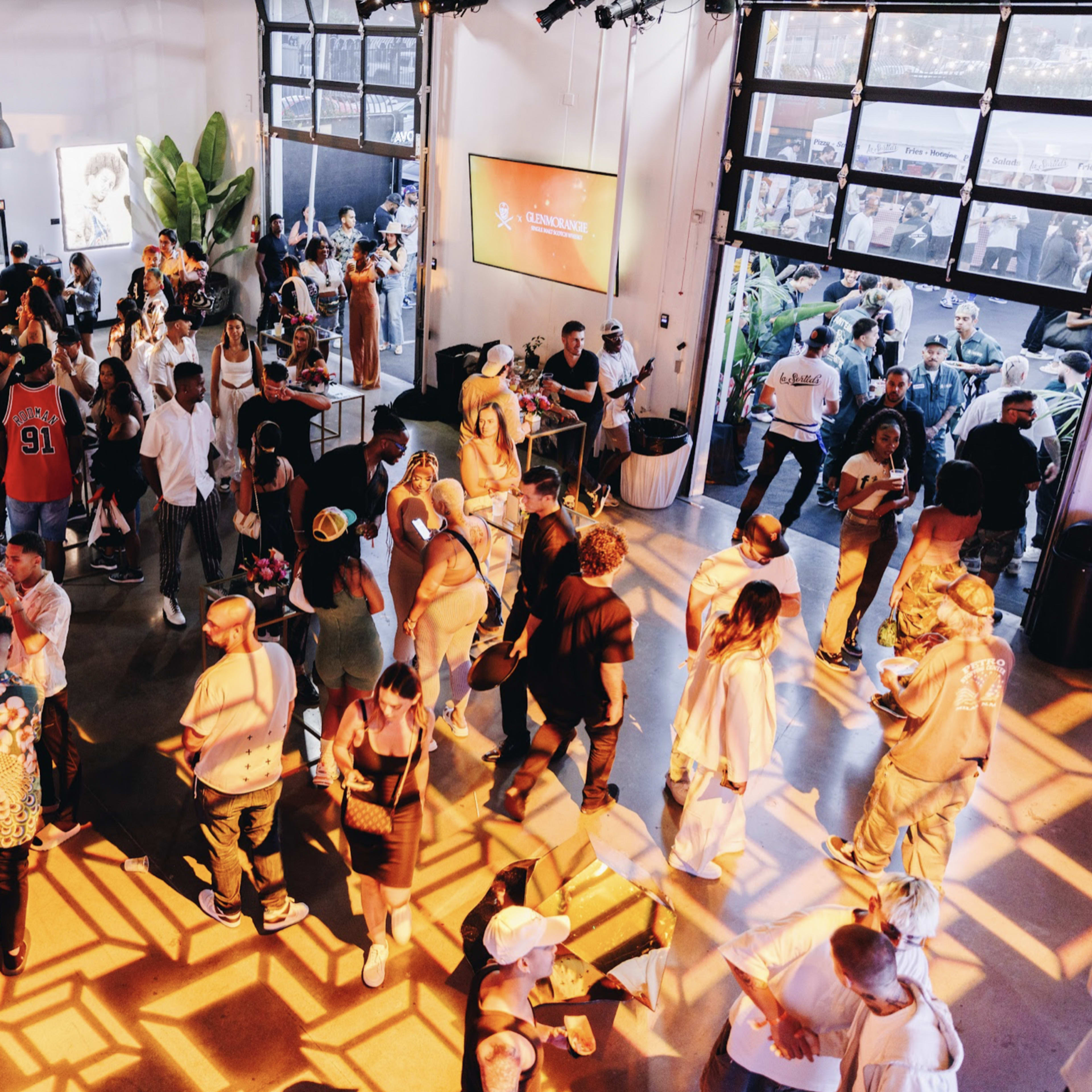 A crowded indoor event space with people mingling and enjoying a vibrant atmosphere, while sunlight casts patterned shadows on the floor.
