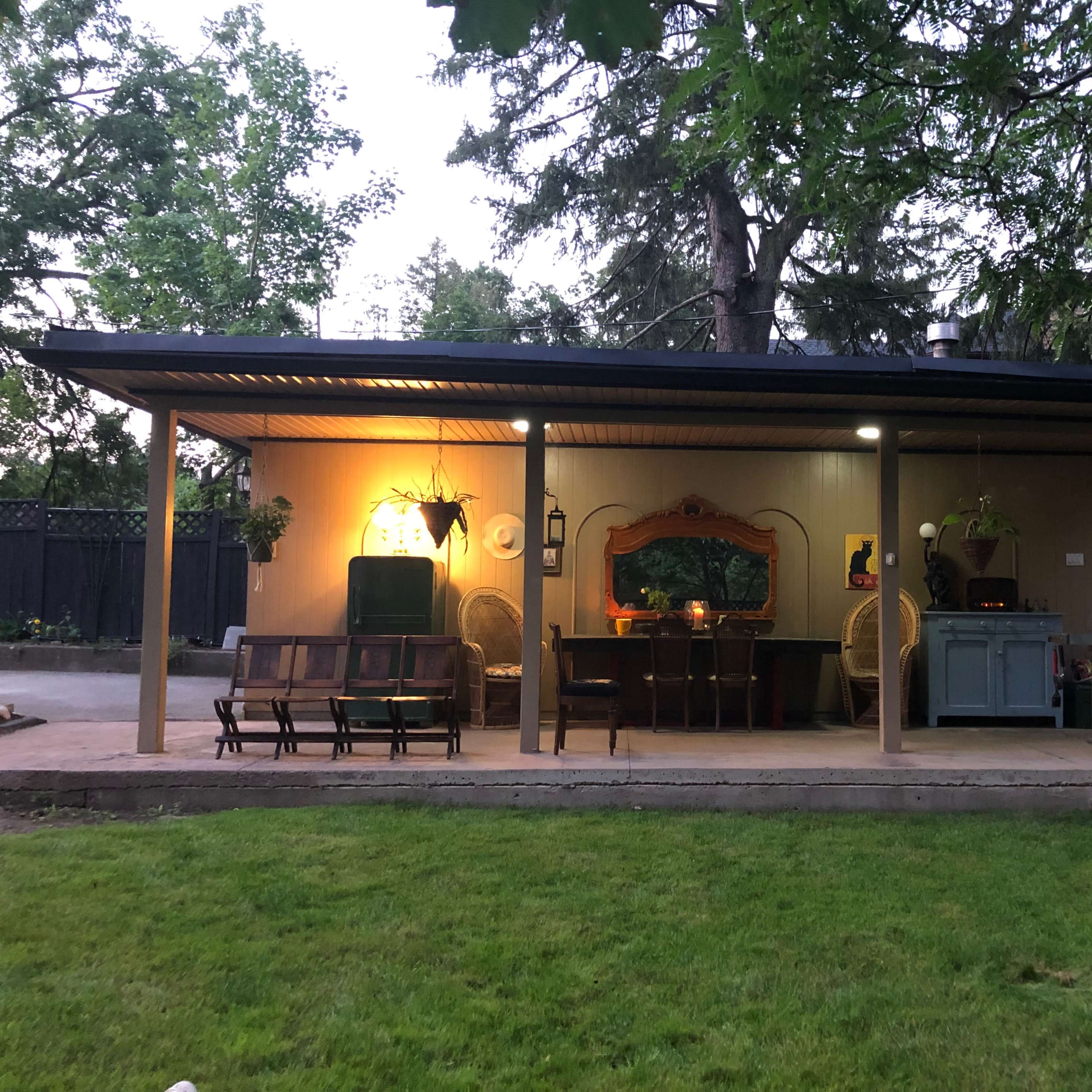 A covered patio with a dining table and chairs, illuminated by warm lights, is surrounded by trees and greenery.