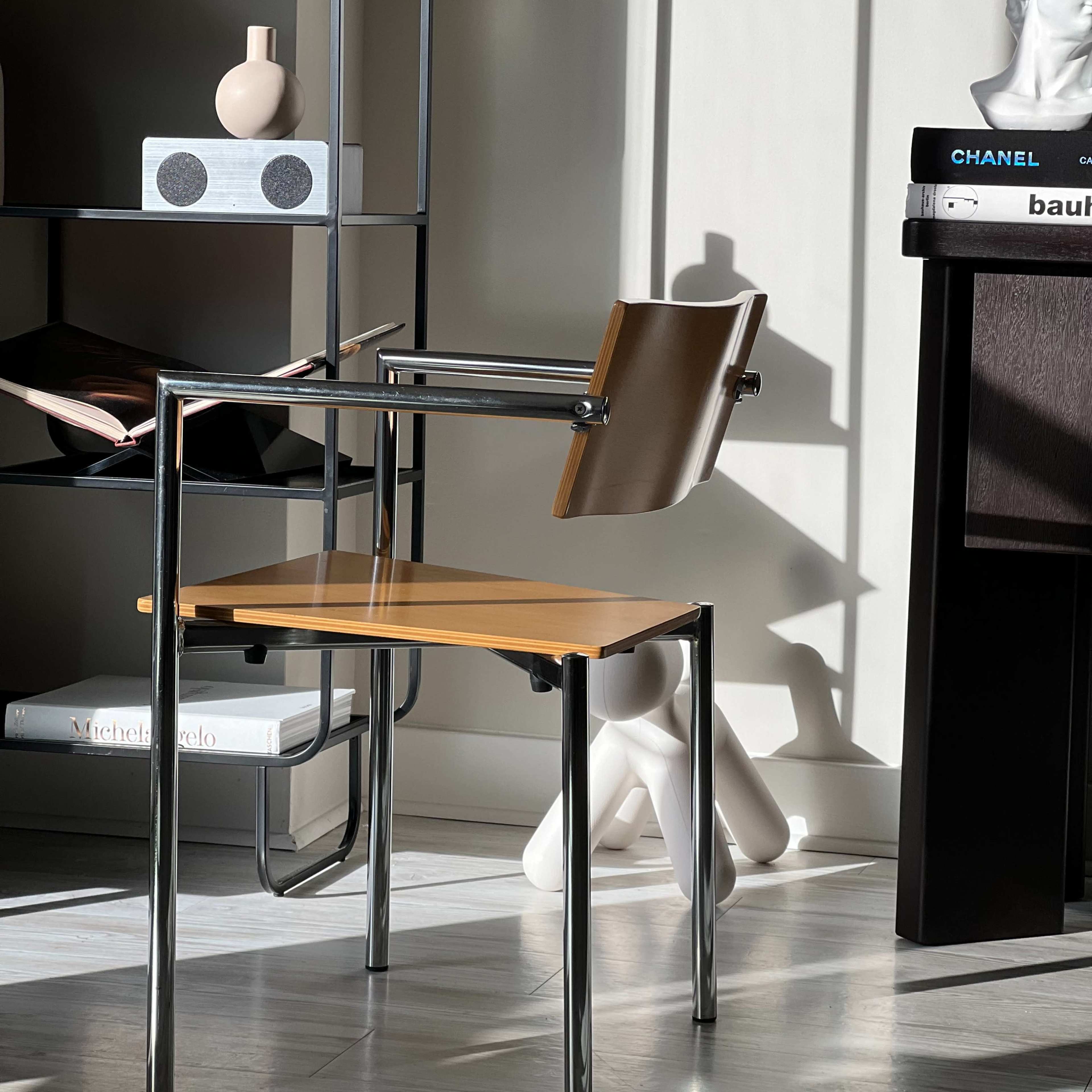 A metal and wood chair sits beside a bookshelf and a decorative figure, illuminated by sunlight casting shadows on the wall.