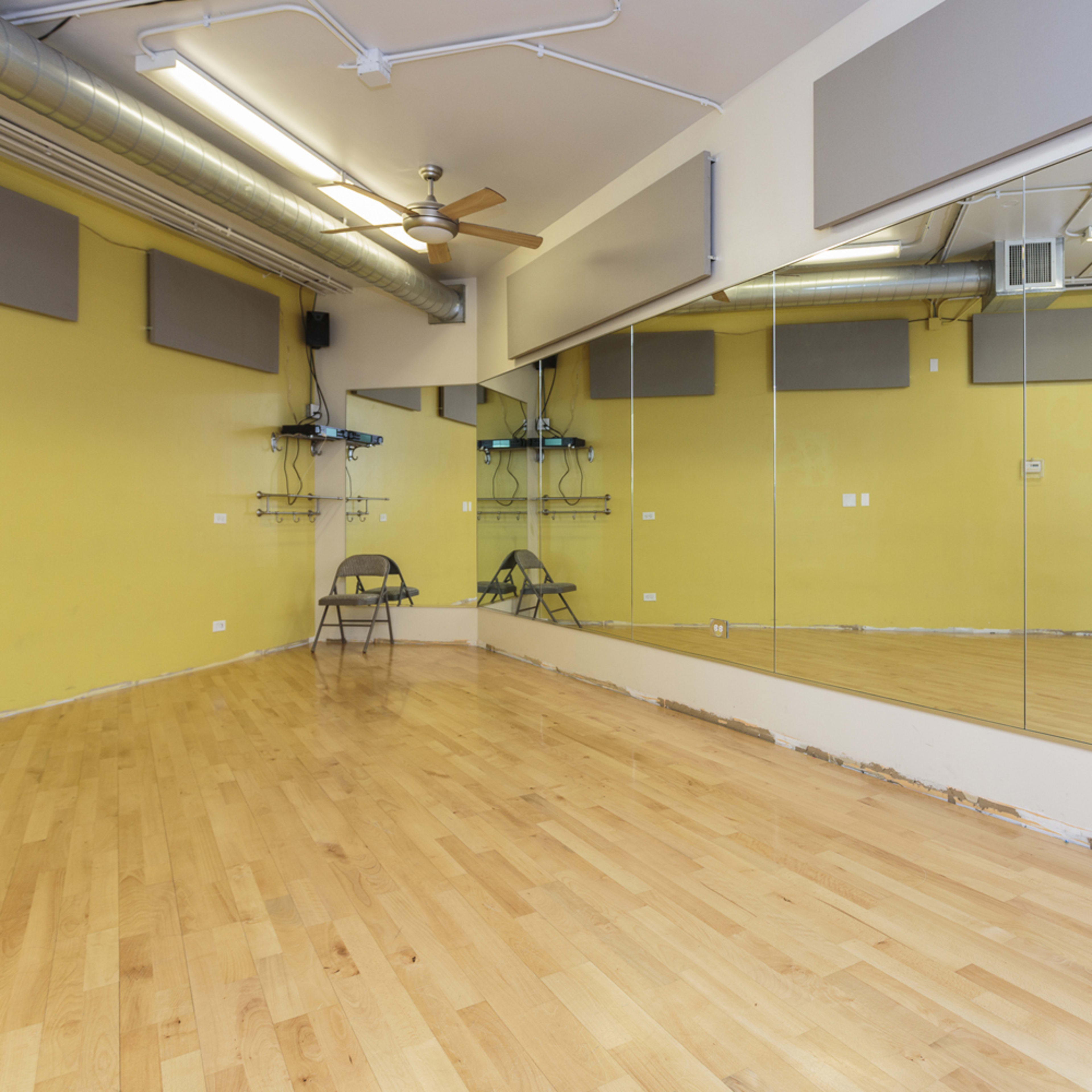 A spacious dance studio with wooden flooring, a large mirror on one wall, and minimal furnishings.