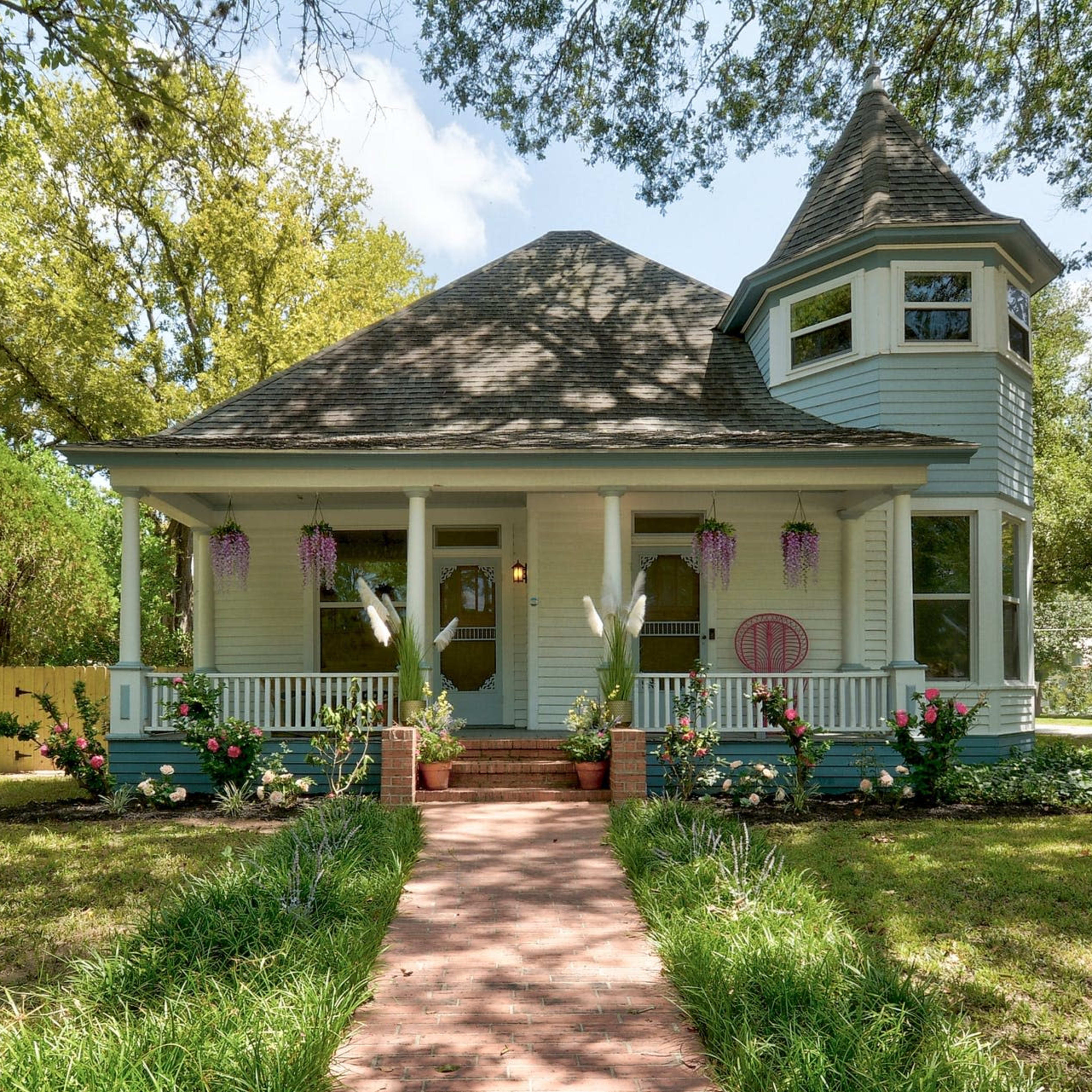 A light blue Victorian-style house with a turret and a front porch, surrounded by green grass and flower beds, features a brick pathway leading to the entrance.