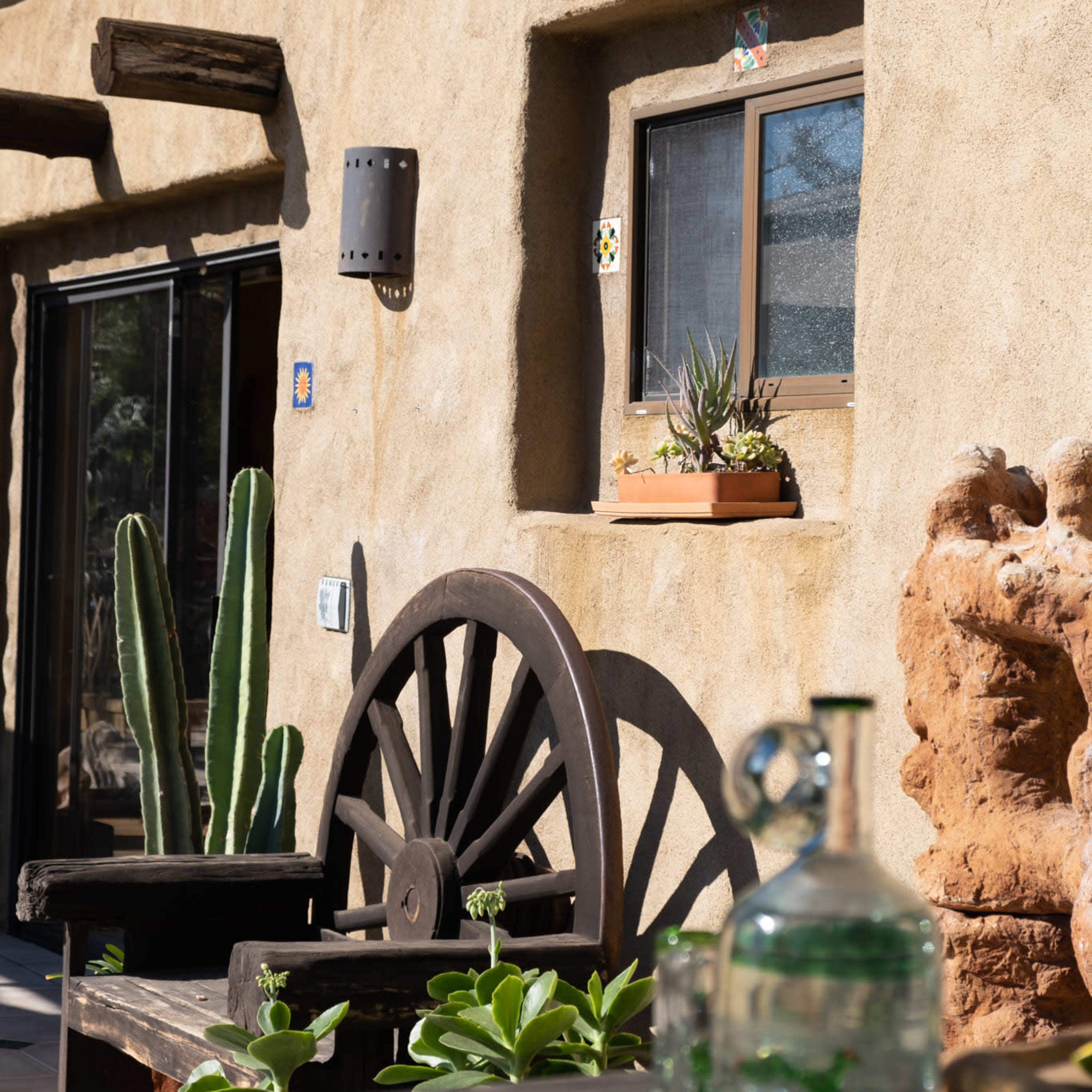 A textured adobe wall with a window adorned with potted plants, alongside a wooden wagon wheel and cacti in a southwestern outdoor setting.