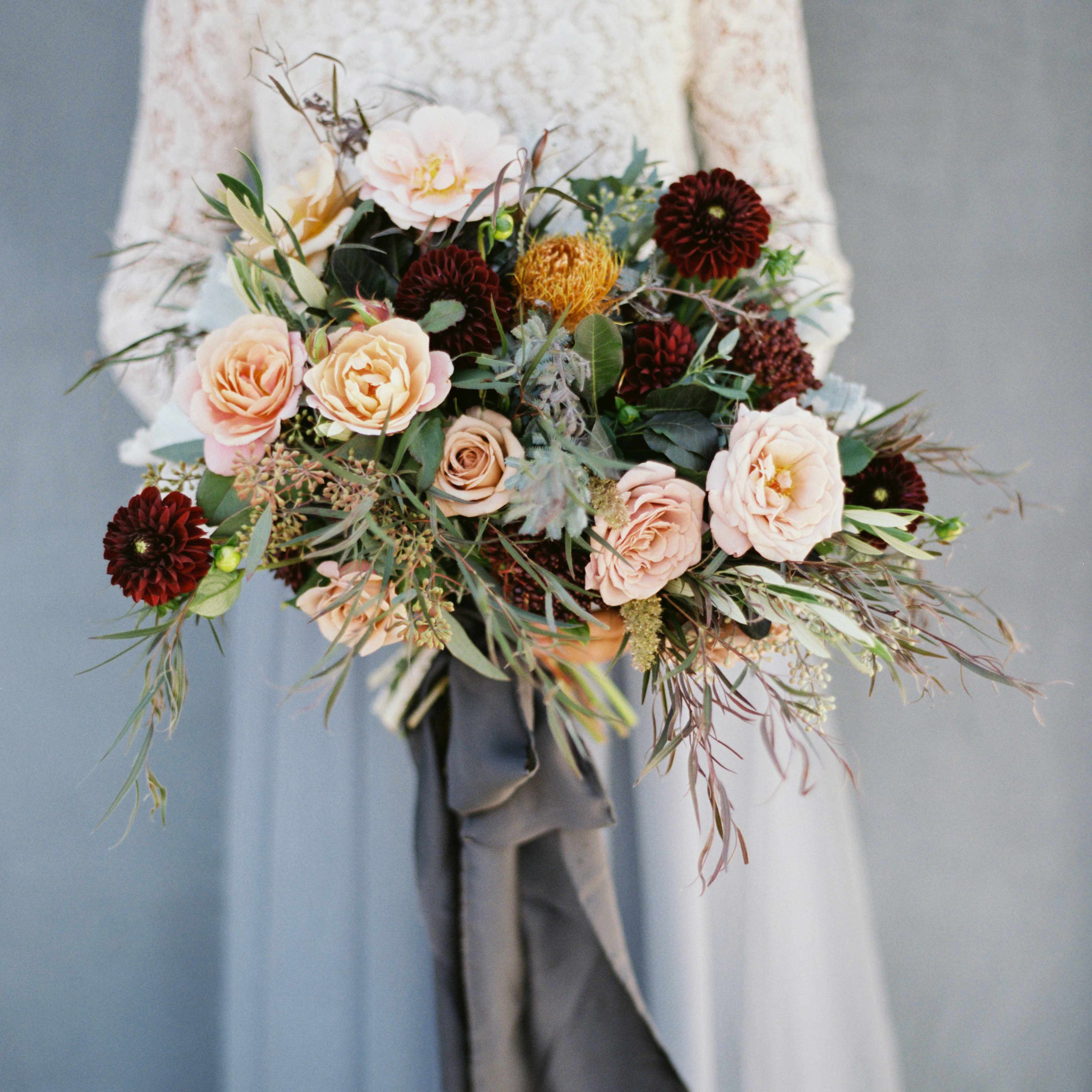 A person holding a bouquet filled with pink roses, brown dahlias, and greenery against a muted background.