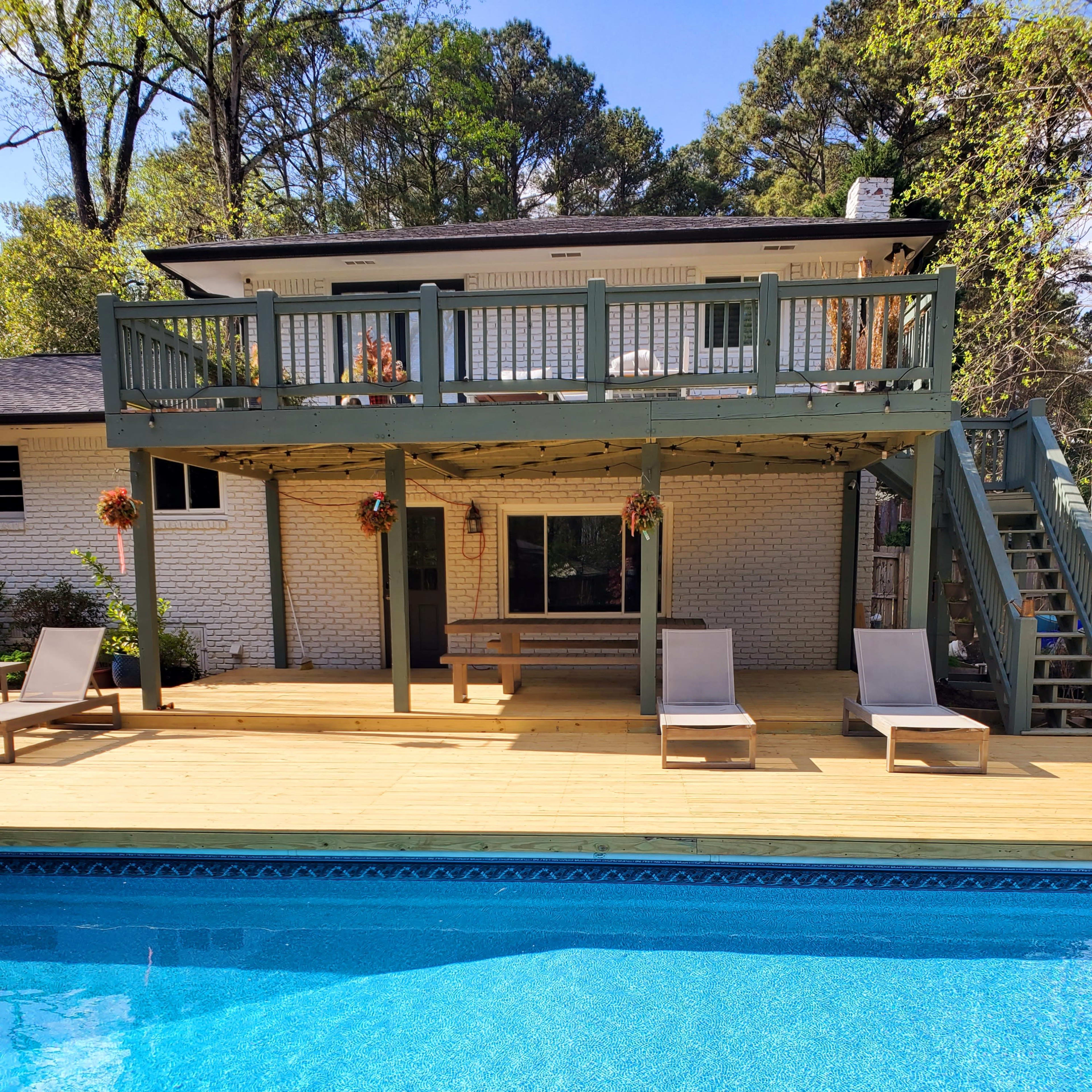 A two-story house with a wooden deck overlooking a swimming pool, featuring lounge chairs beside the pool and a staircase leading to the upper level.