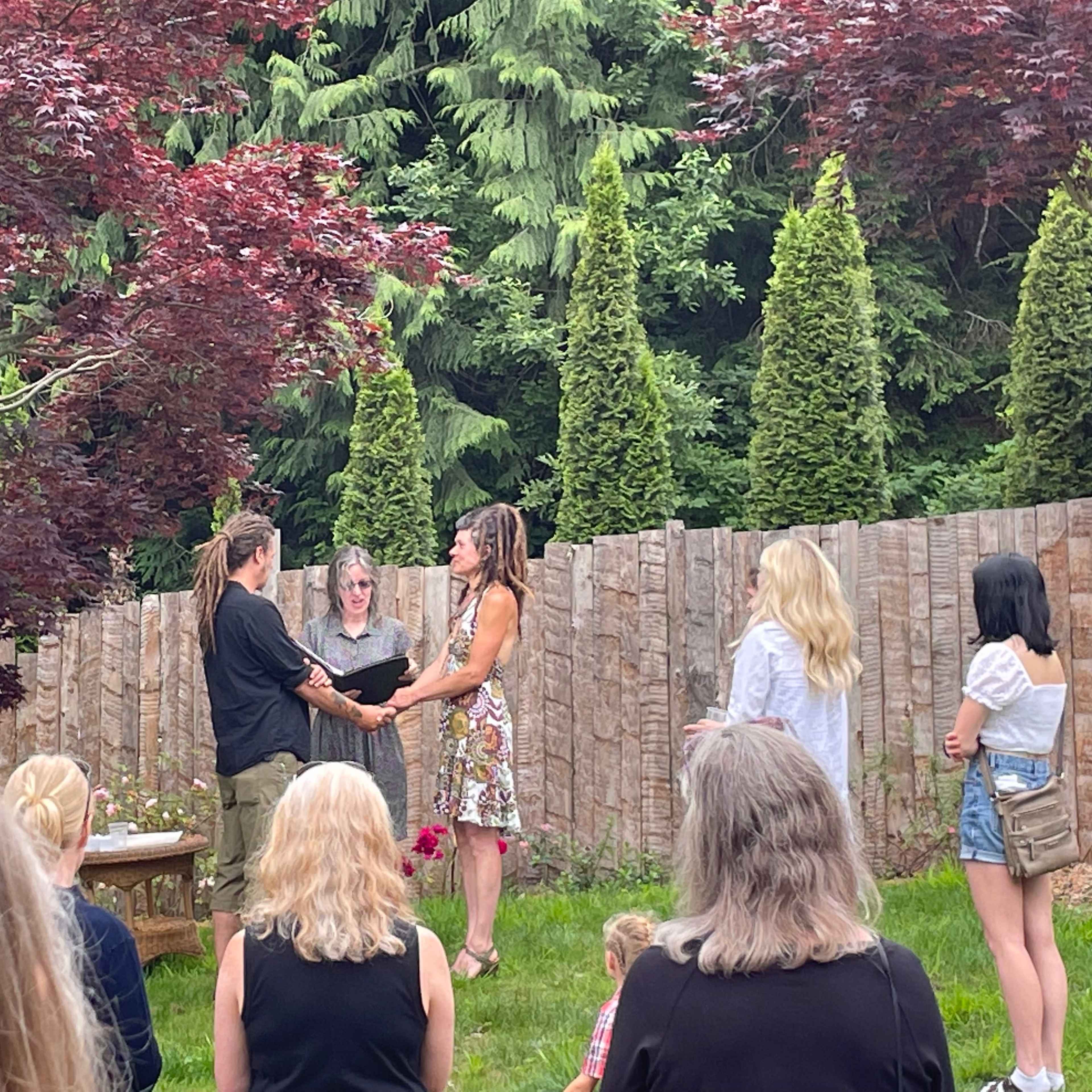 A couple stands hand in hand under a canopy of trees while a small group of people observes them in a backyard setting.