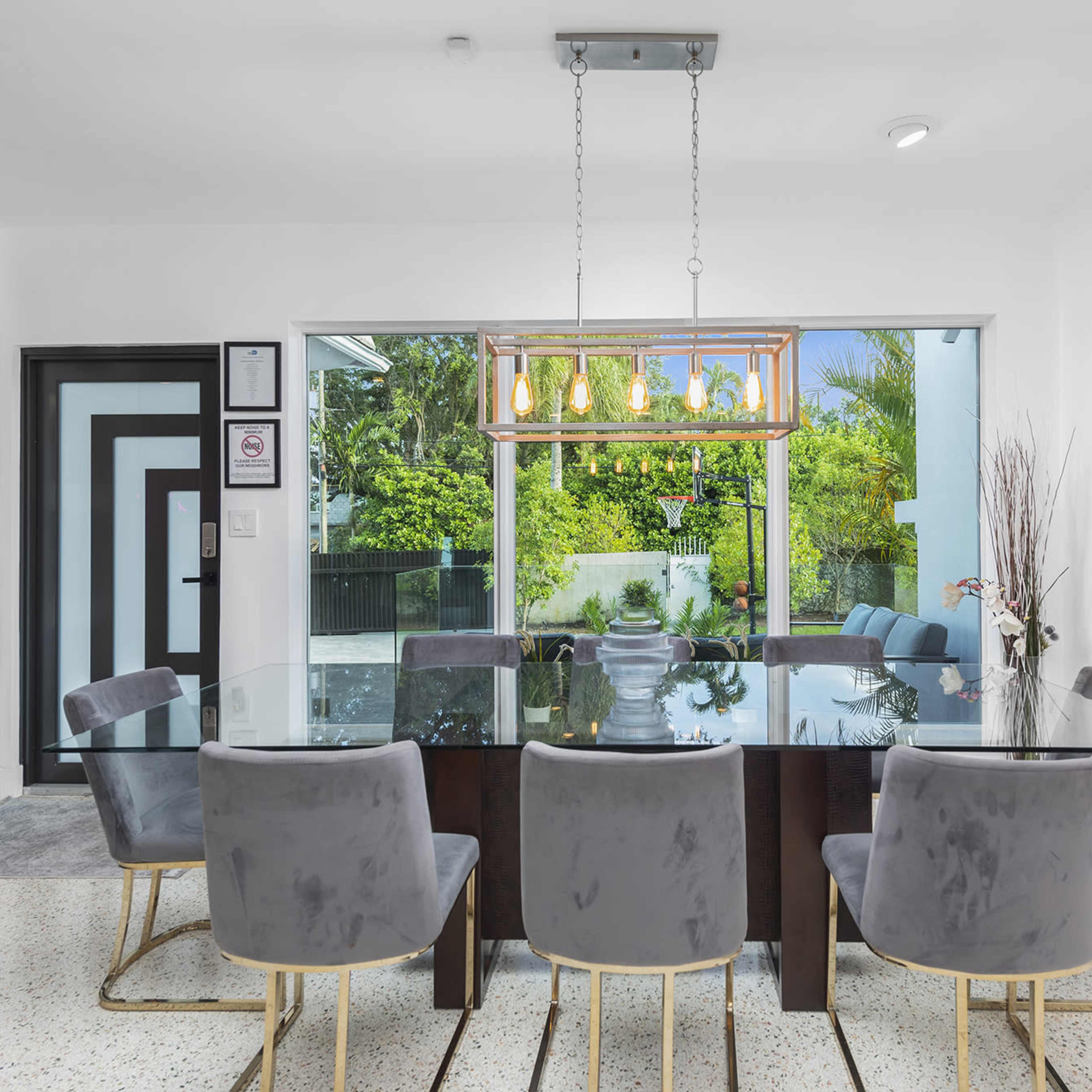 A modern dining room features a glass table surrounded by six upholstered chairs, with a large window showcasing a view of greenery outside.