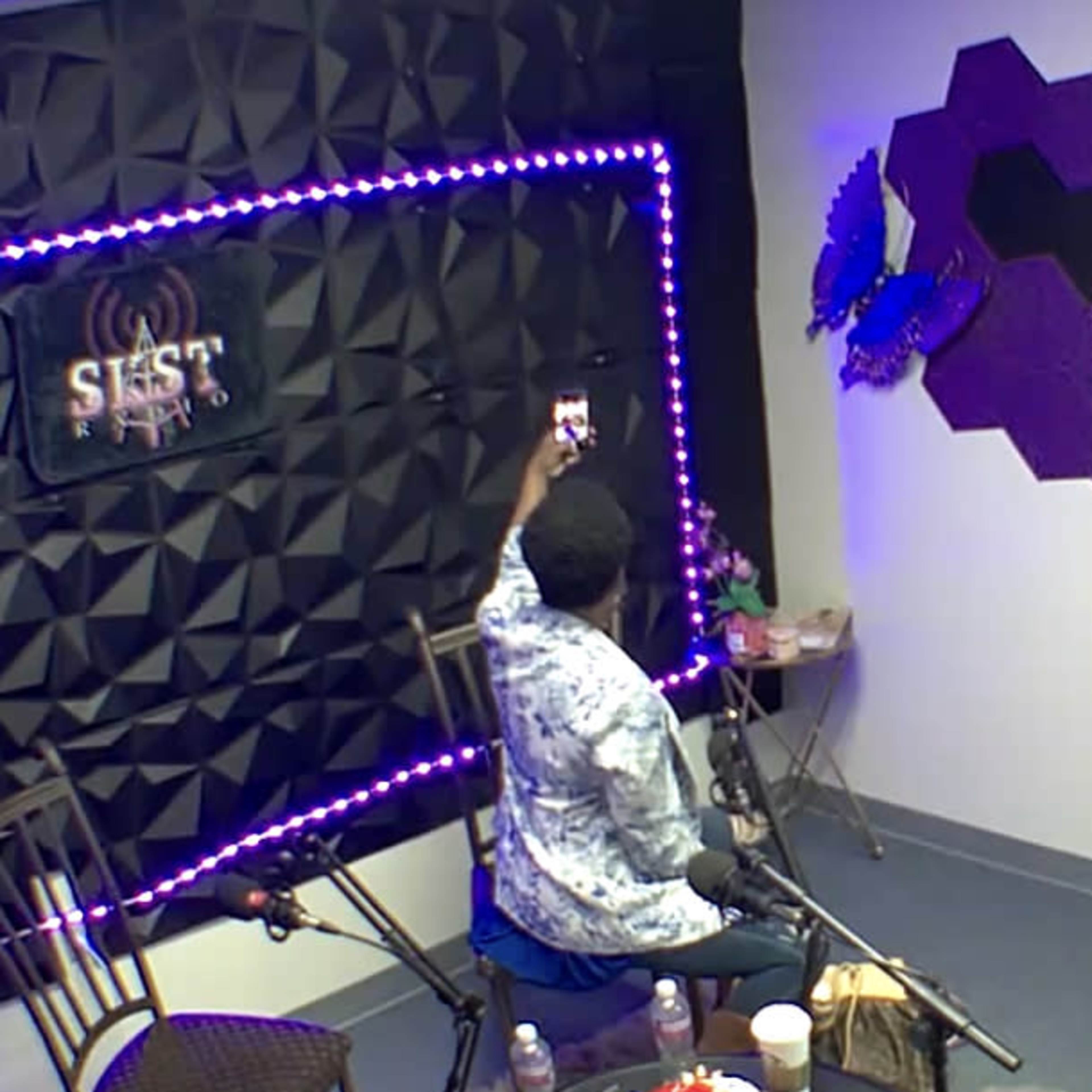 A person sits in a studio, holding a phone, with a backdrop of dim lighting and decorative elements on the wall.