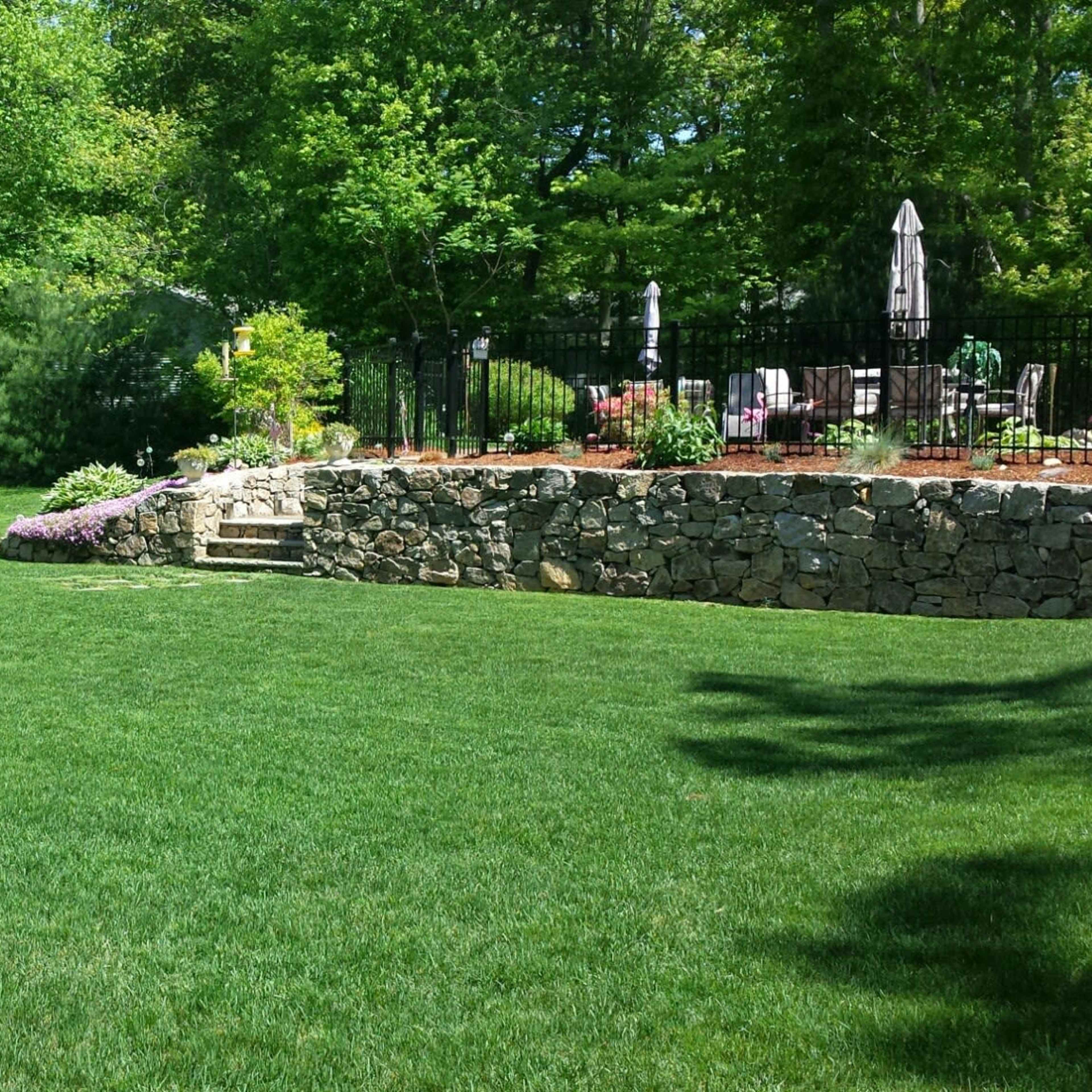 A neatly manicured lawn with a stone wall and landscaped garden beds is surrounded by trees and a house.