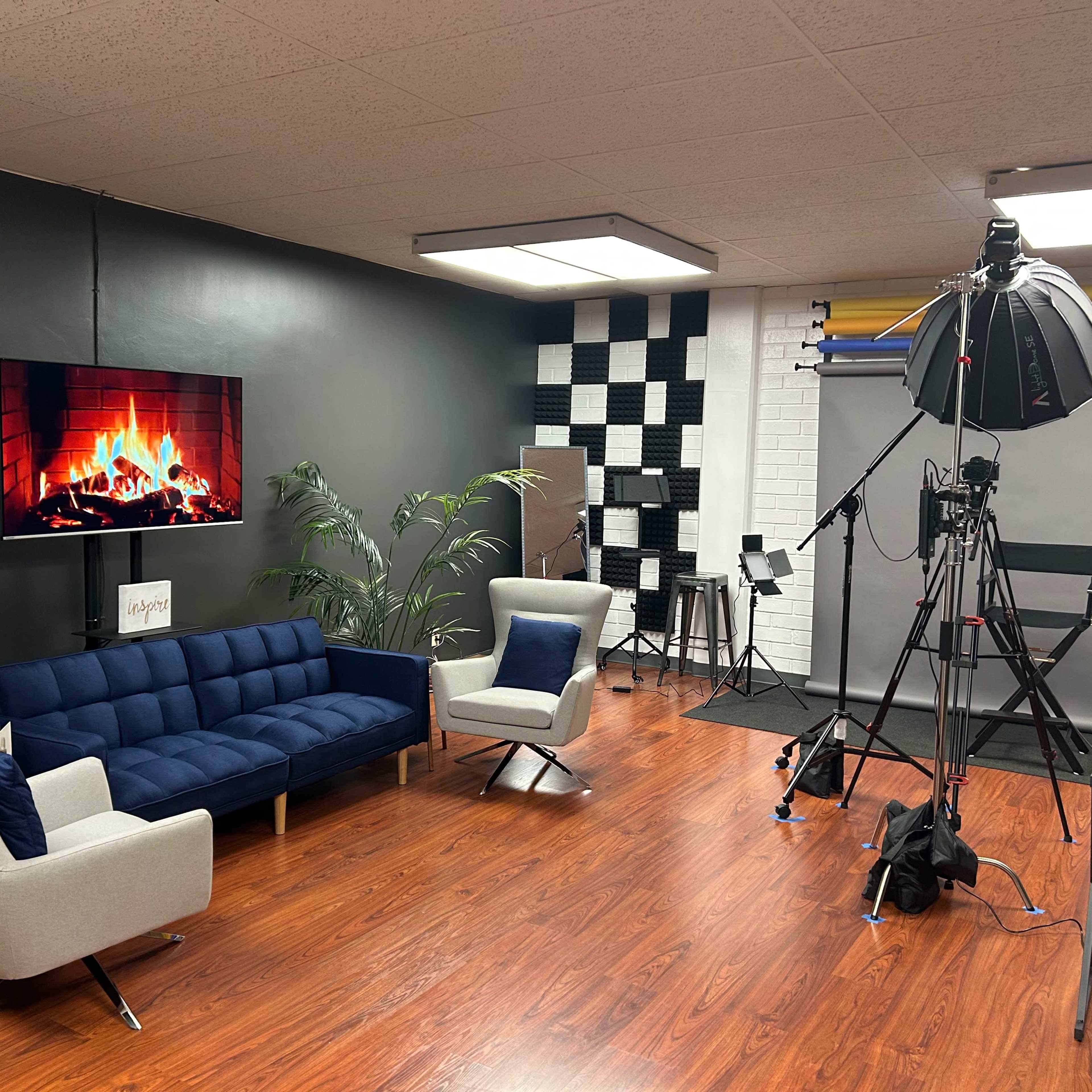 A modern studio setup featuring a blue couch, an artificial fireplace display on the wall, and various filming equipment including lighting and tripods.