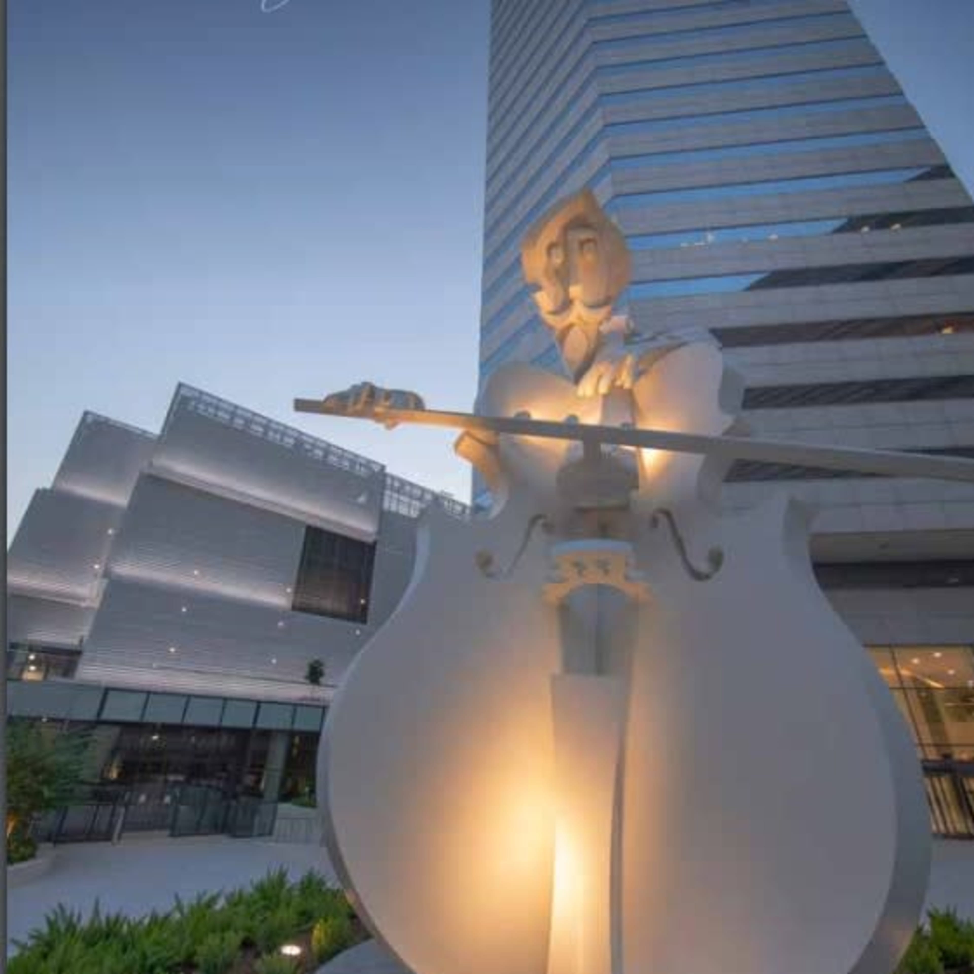 A large, illuminated statue of a musician playing a cello stands in front of a modern building.