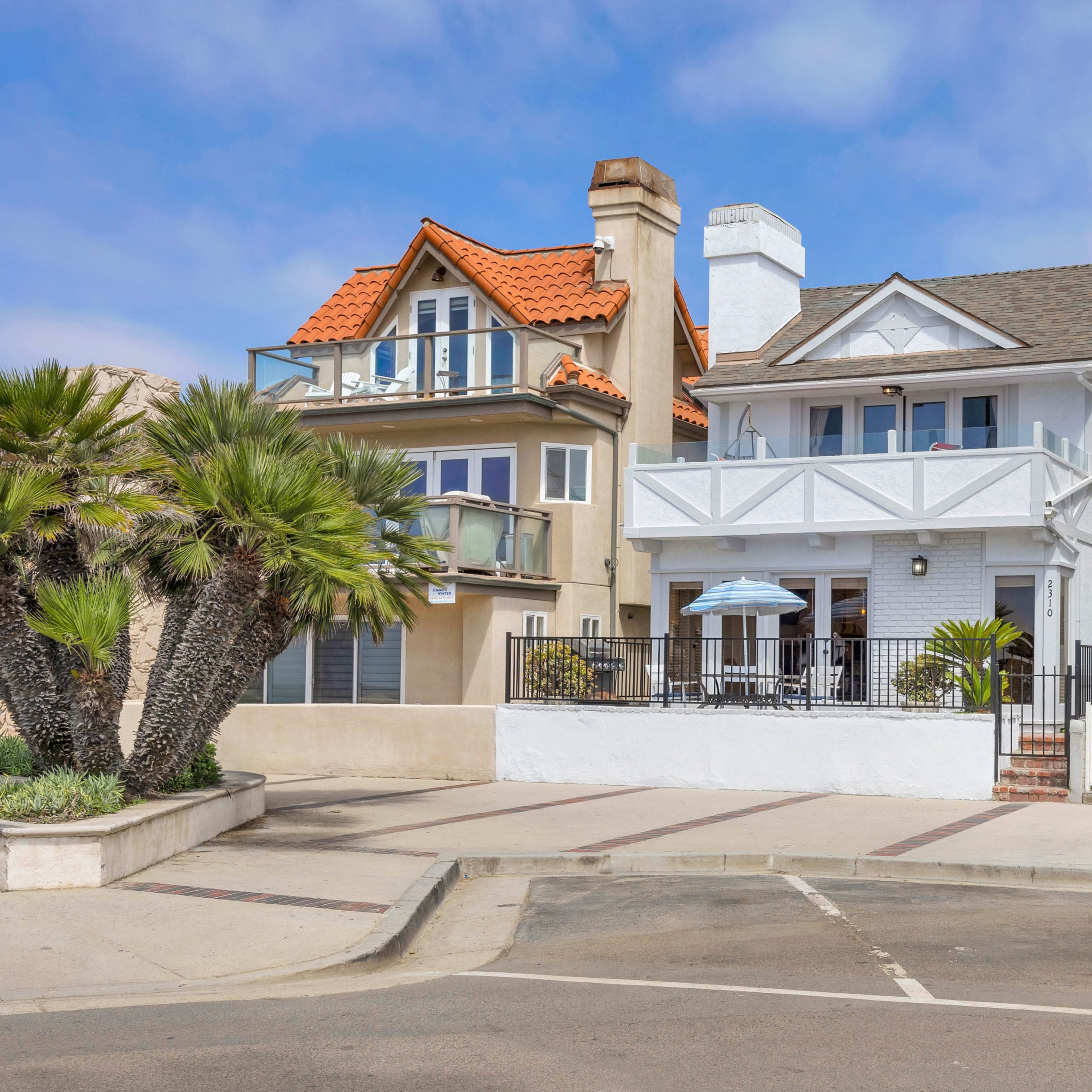 A row of beachfront houses, including a white two-story home with a front porch, flanked by a house with a red-tiled roof and a modern glass-faced property.