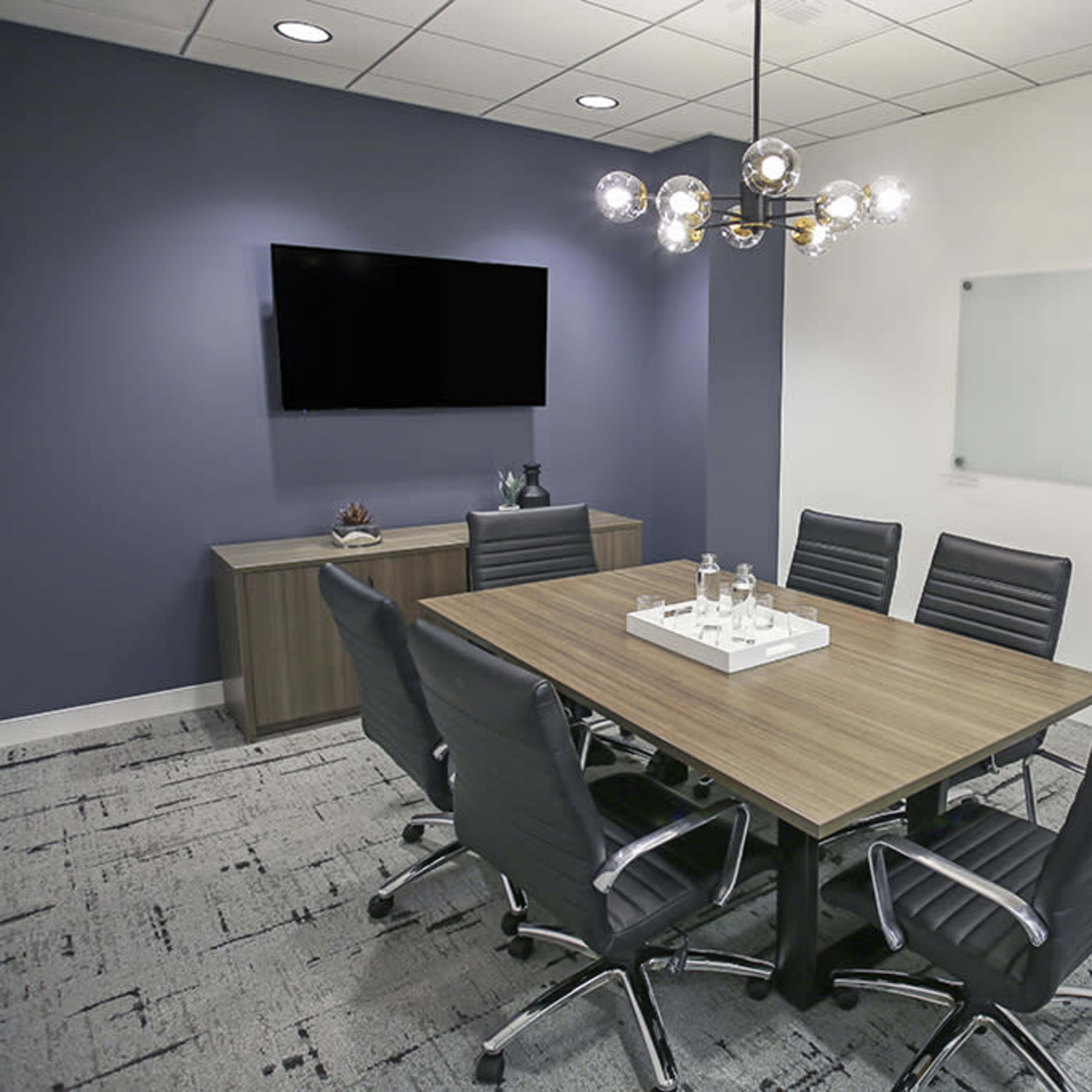 A modern conference room features a wooden table surrounded by six black chairs, a television mounted on the wall, and a glass whiteboard.